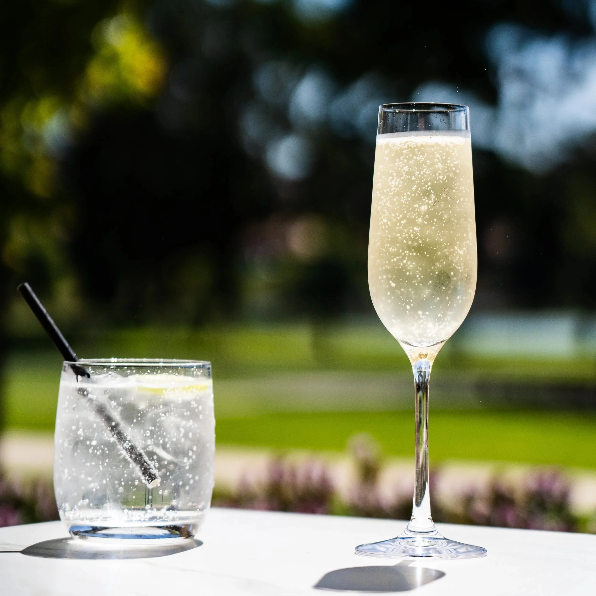 A glass of clear bubbly drink with a straw and a lemon slice on a table outdoors, with a blurred green background and a tall glass of champagne nearby.