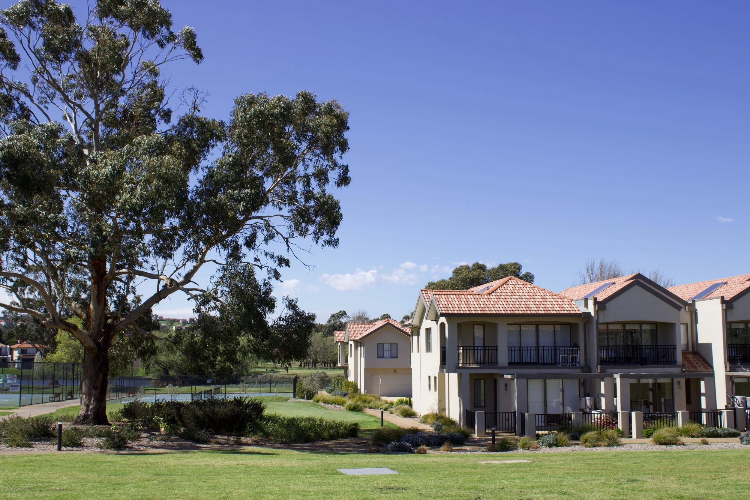 Residential complex with multi-story townhouses featuring red-tiled roofs, balconies, and front gardens, situated in a landscaped park with green lawns, trees, and a tennis court, under a clear blue sky.