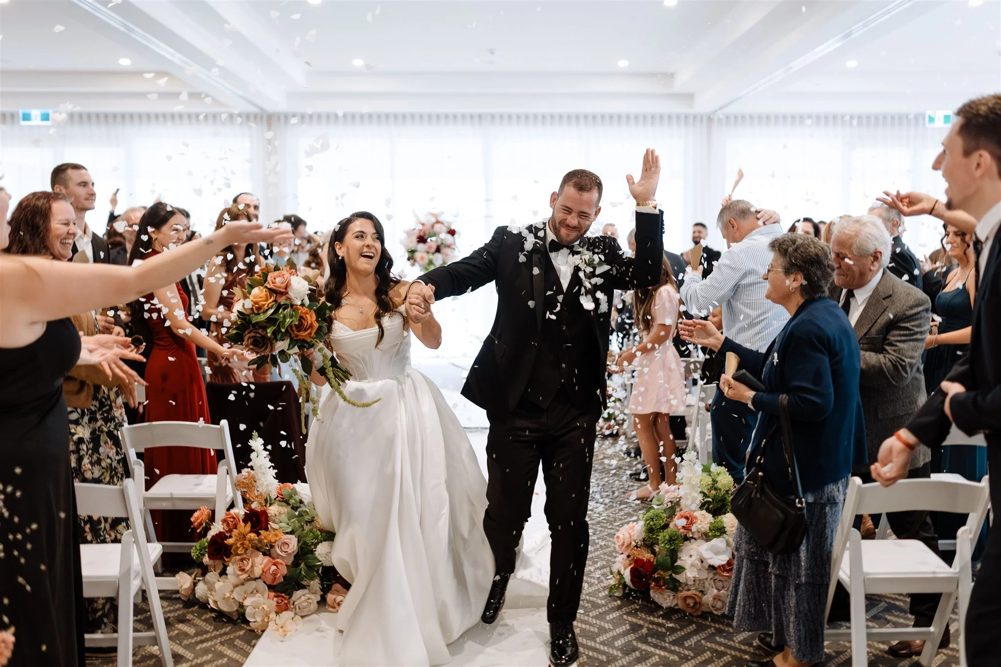 Bride and groom entering wedding reception, holding hands and smiling, surrounded by guests throwing confetti, floral arrangements, and smiling.