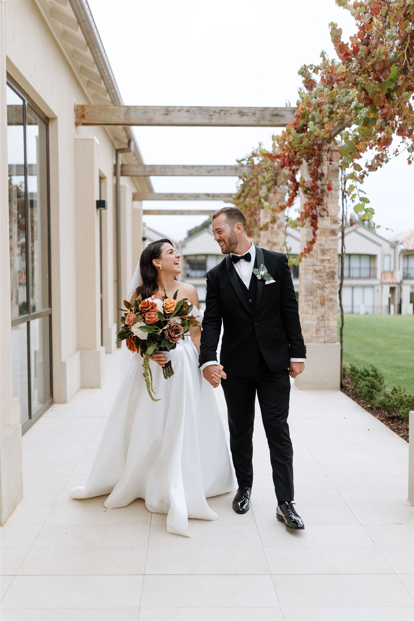 A newlywed couple walking hand in hand outside, smiling at each other. The bride is holding a bouquet of flowers, wearing a white wedding gown, and the groom is in a black tuxedo with a bow tie. They are in an outdoor area with green grass, autumn-co
