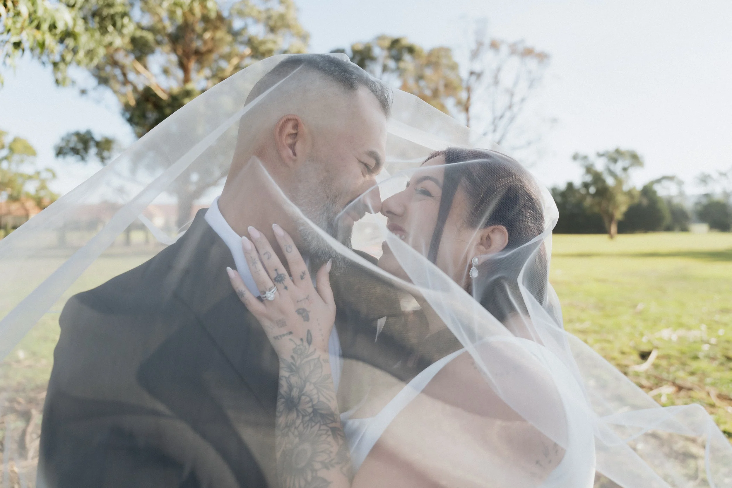 A bride and groom in wedding attire under a veil, smiling and leaning close to each other outdoors with trees and a grassy field in the background.