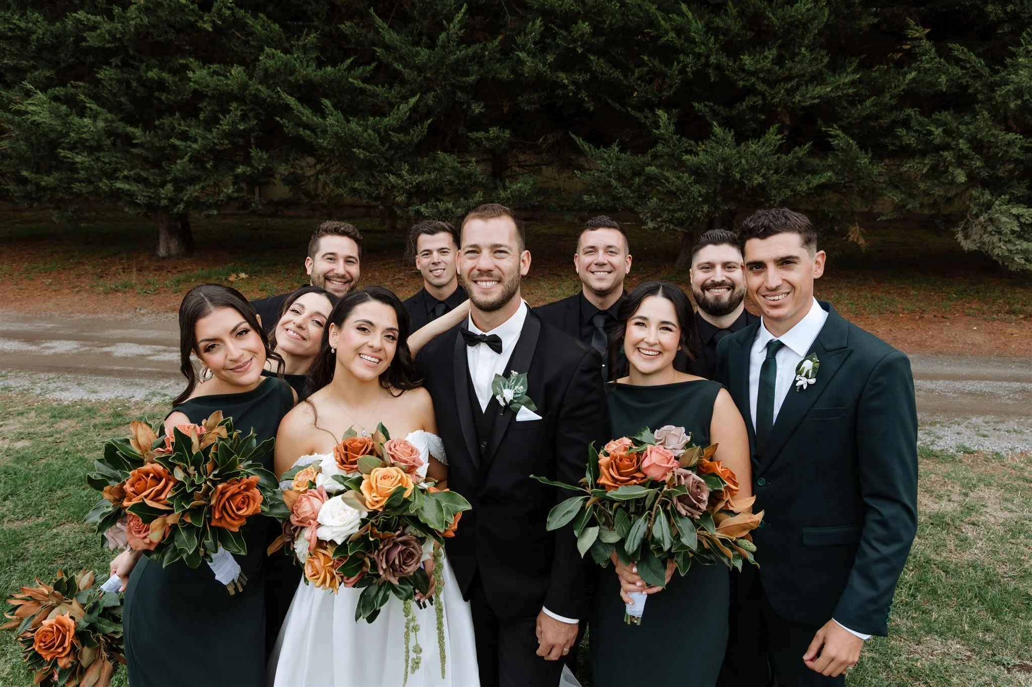A wedding party of ten people outdoors, with the bride and groom in the center holding bouquets, surrounded by bridesmaids and groomsmen, all smiling in front of green trees.