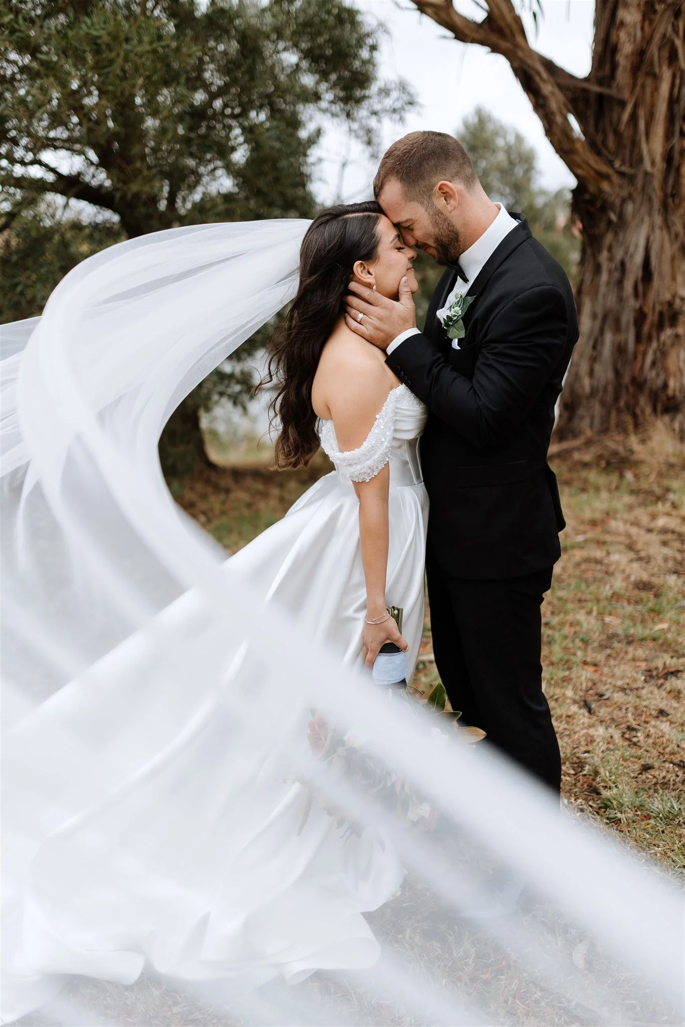 A bride and groom sharing a romantic moment outdoors, with the bride's veil flowing in the wind and holding a bouquet, surrounded by trees.