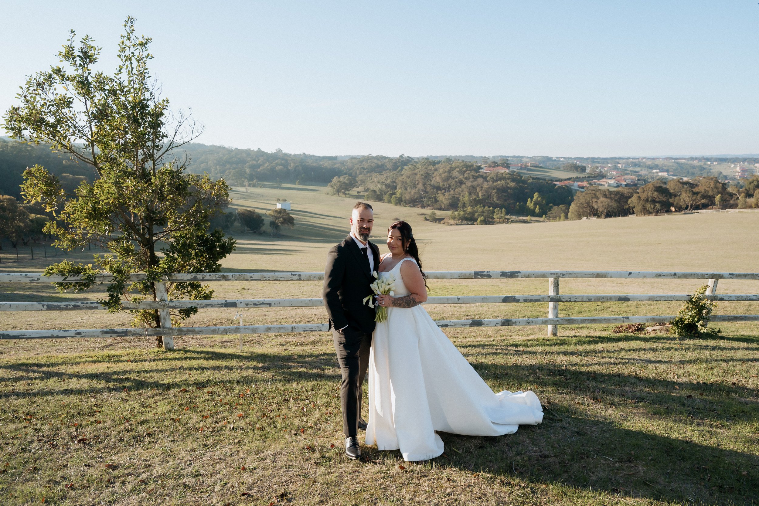 A newlywed couple posing outdoors in a rural landscape. The groom, dressed in a black suit, and the bride, wearing a white wedding gown, stand in front of a white wooden fence with rolling hills and trees in the background. The bride holds a bouquet 