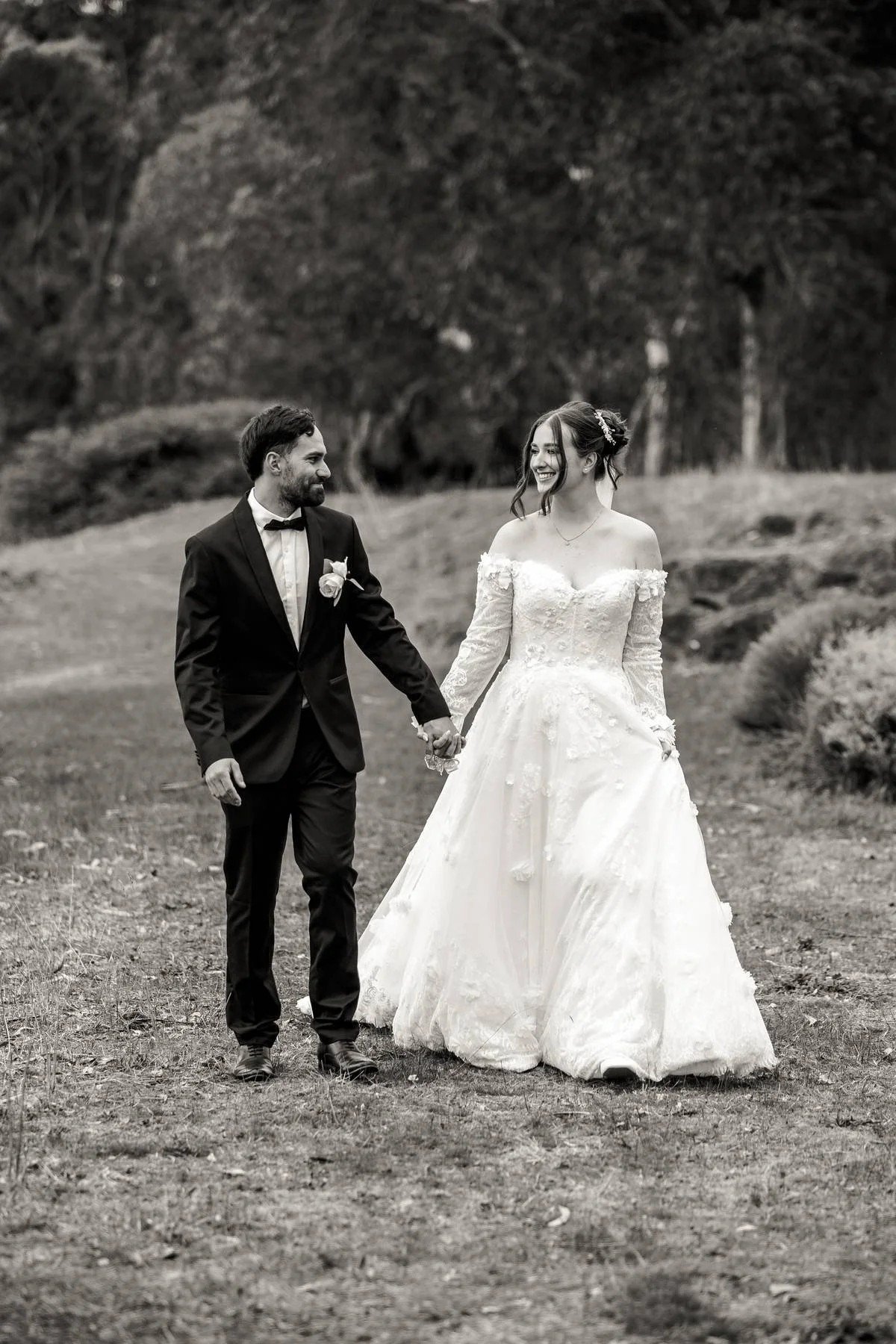 A bride and groom holding hands and walking outdoors in a natural setting, smiling at each other.