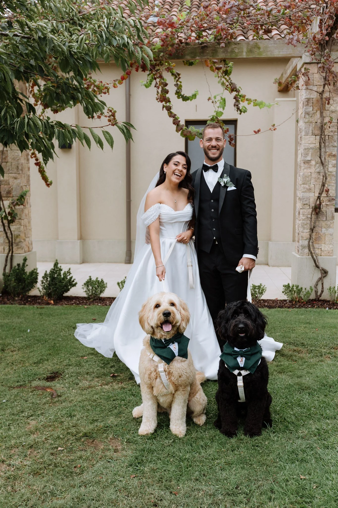 A bride and groom in wedding attire standing on grass, smiling, with two dogs wearing green bow ties, in front of a building with brick and stone walls, and greenery.