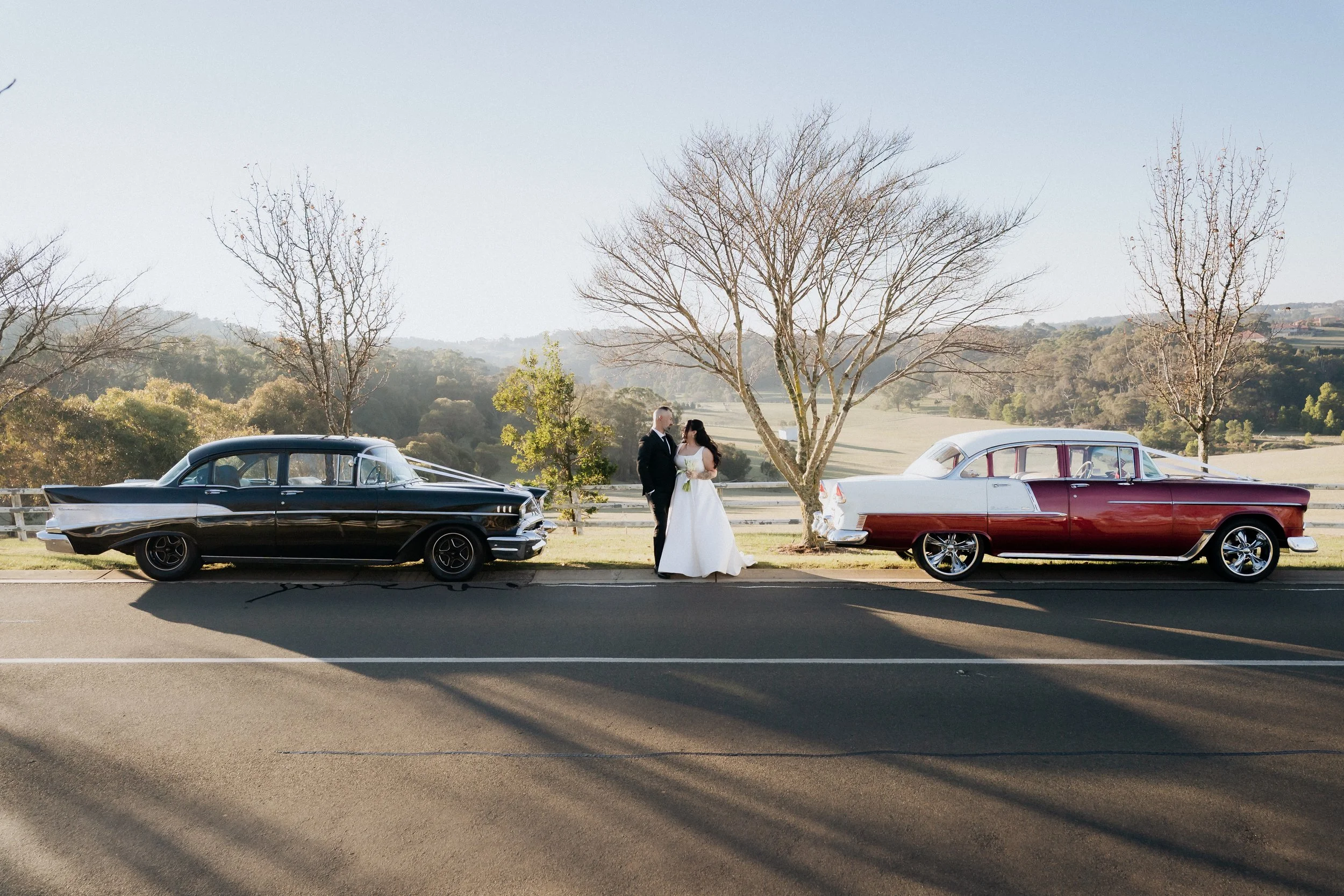 A bride and groom standing between two vintage cars on a rural road with a scenic background of trees and rolling hills.
