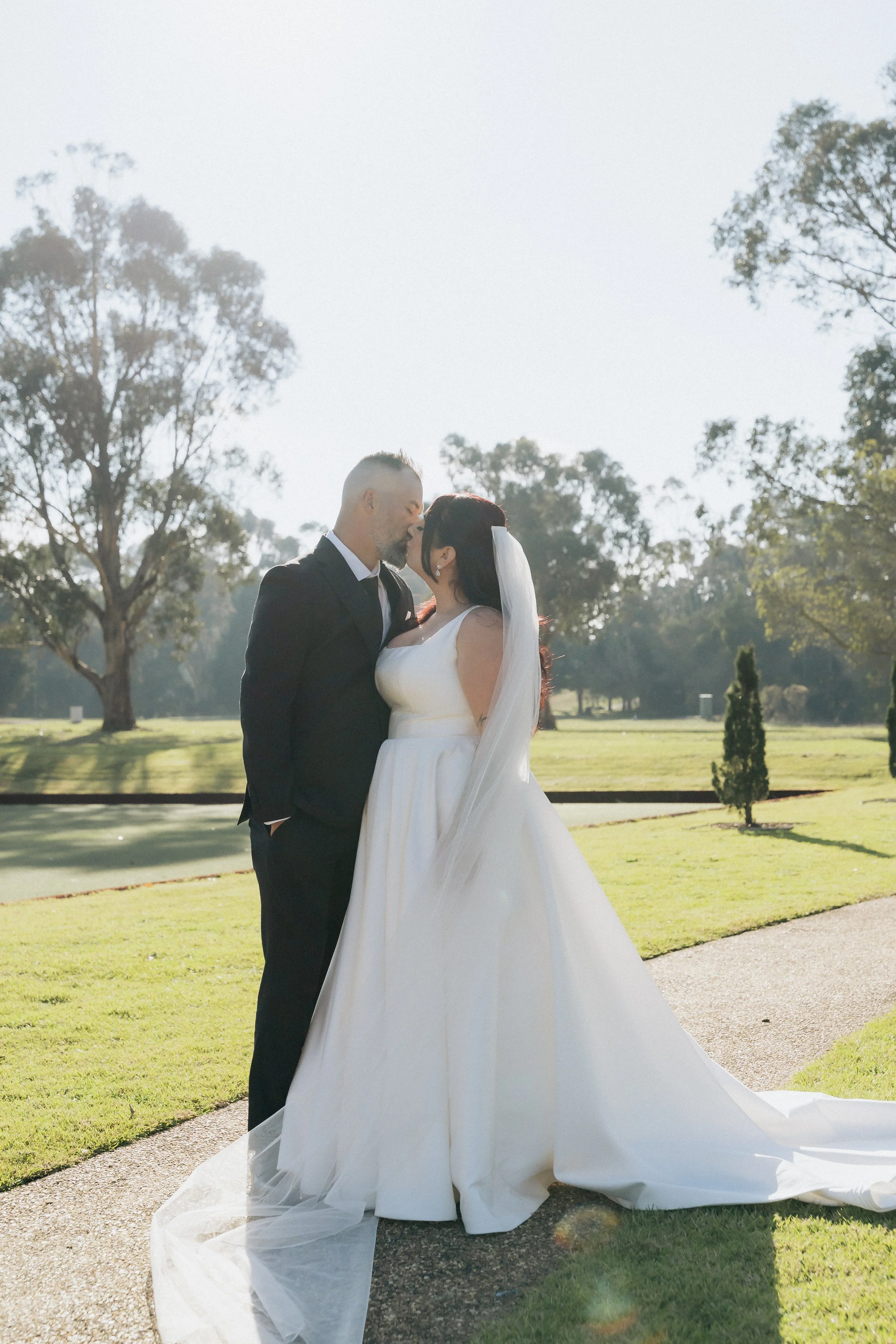 A bride and groom sharing a kiss outdoors in a park on their wedding day, dressed in wedding attire with trees and a lawn in the background.