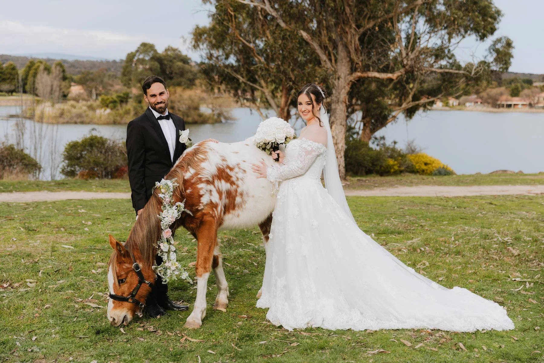 A bride and groom standing outdoors with a small brown and white horse decorated with flowers, near a lake or pond, with trees and a cloudy sky in the background.