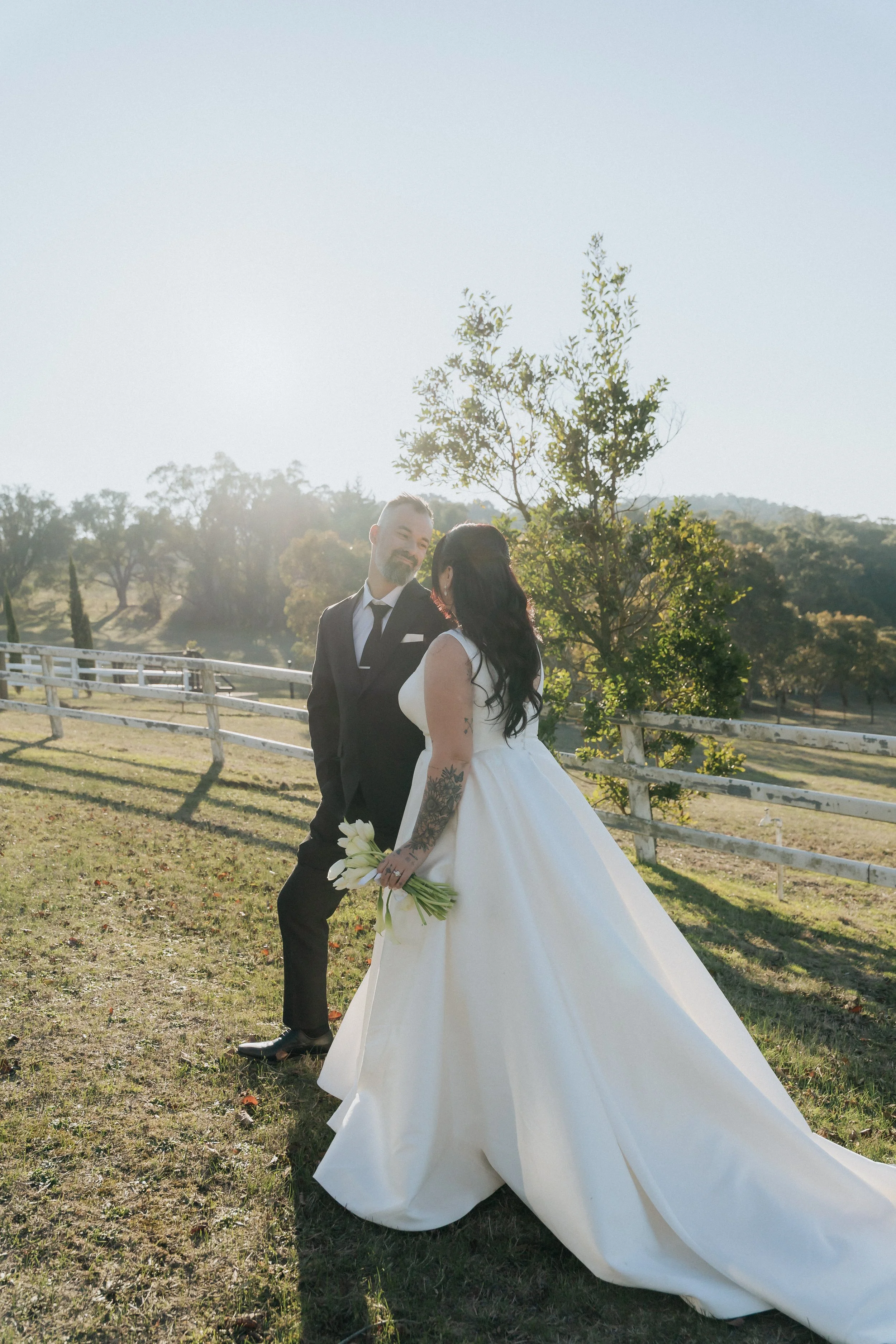 A bride in a white gown holding flowers and a groom in a black suit outside on a sunny day, with a wooden fence and trees in the background.