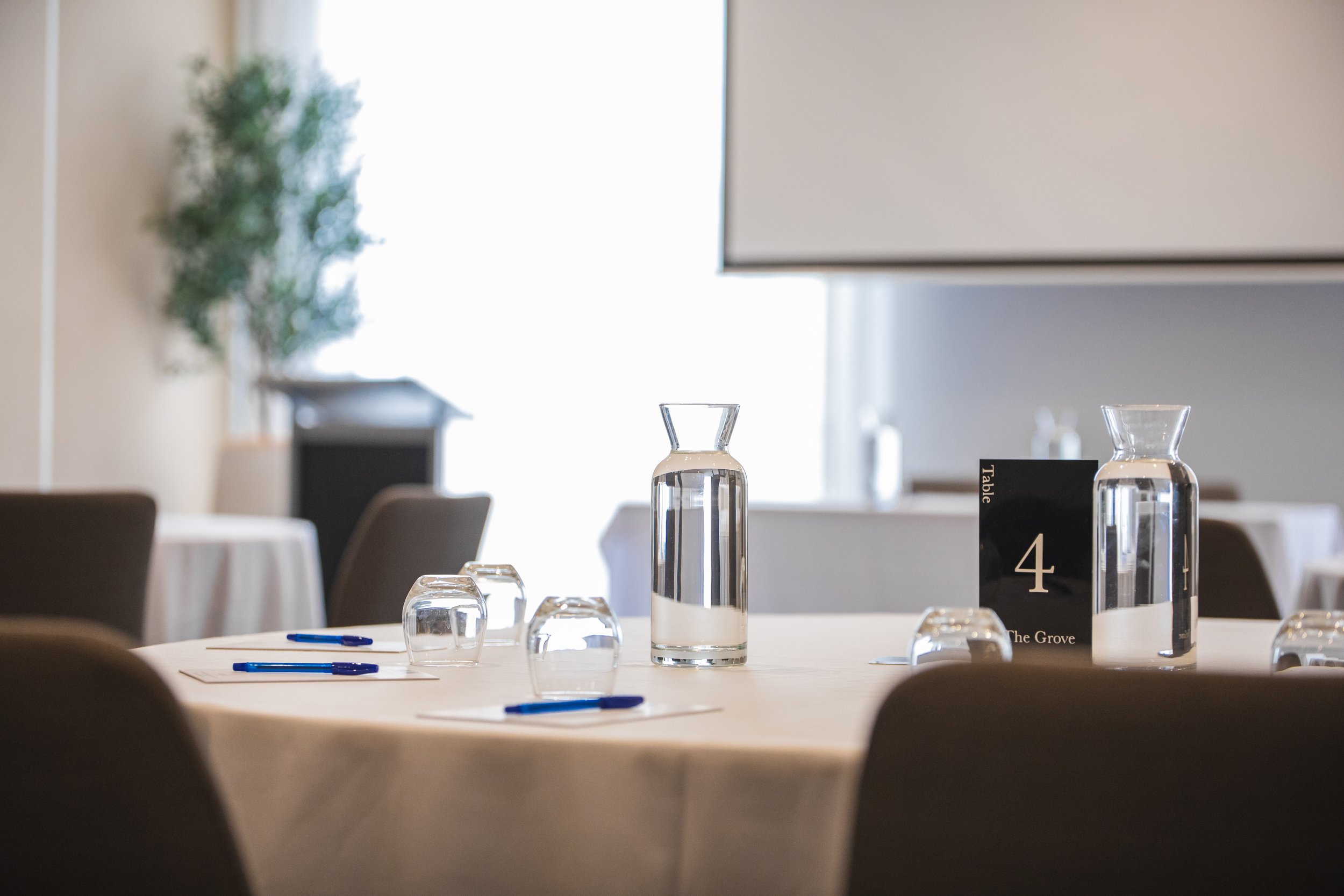 A round banquet table set for a meal with a white tablecloth, glass water pitchers, upside-down glasses, blue pens, and a table number card reading '4' in a formal dining room.