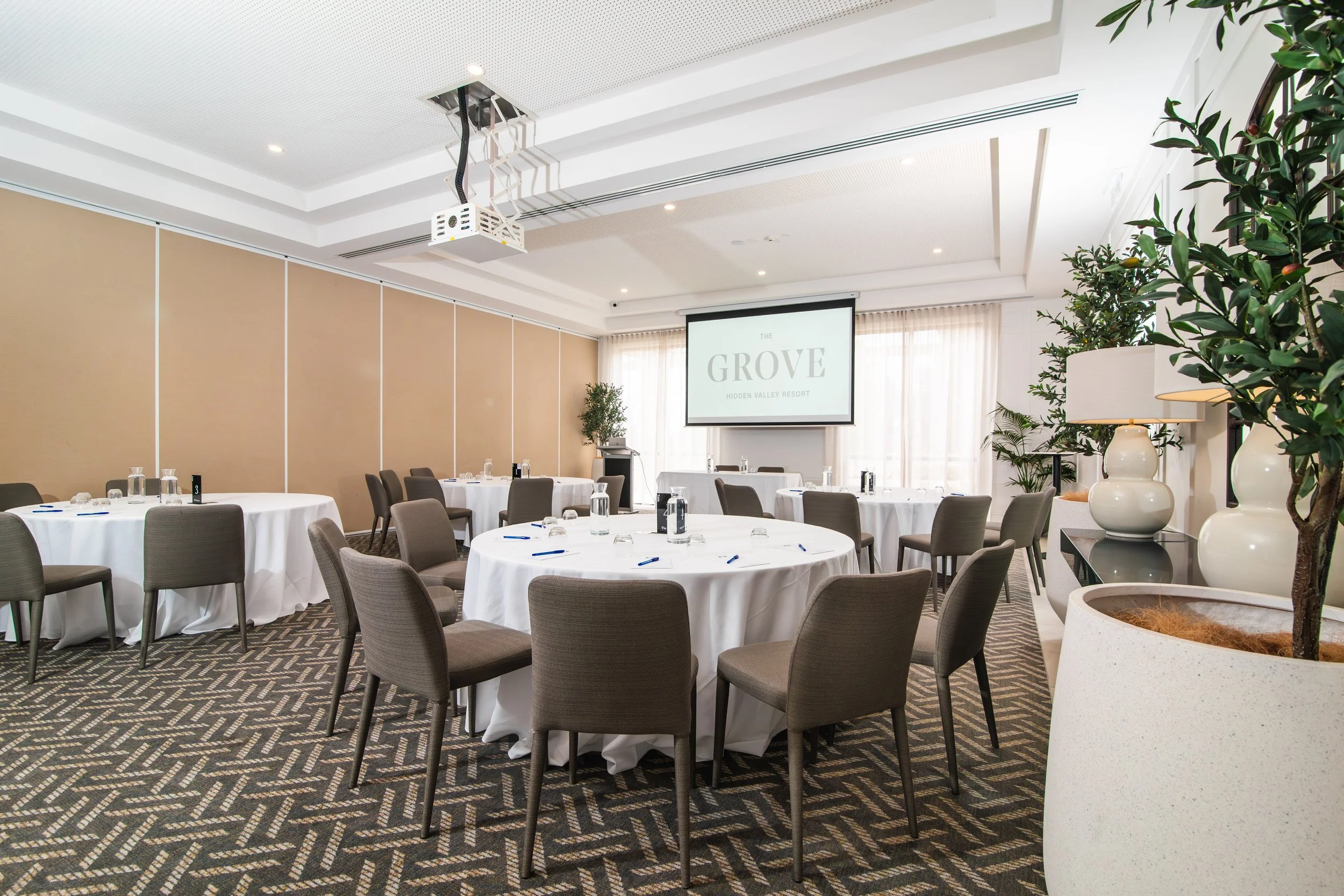 A conference room set up with round tables covered in white tablecloths, each with chairs, water glasses, notepads, and pens. There is a large screen at the front displaying 'The Grove Hidden Valley Resort.' The room has beige walls, curtains, and de