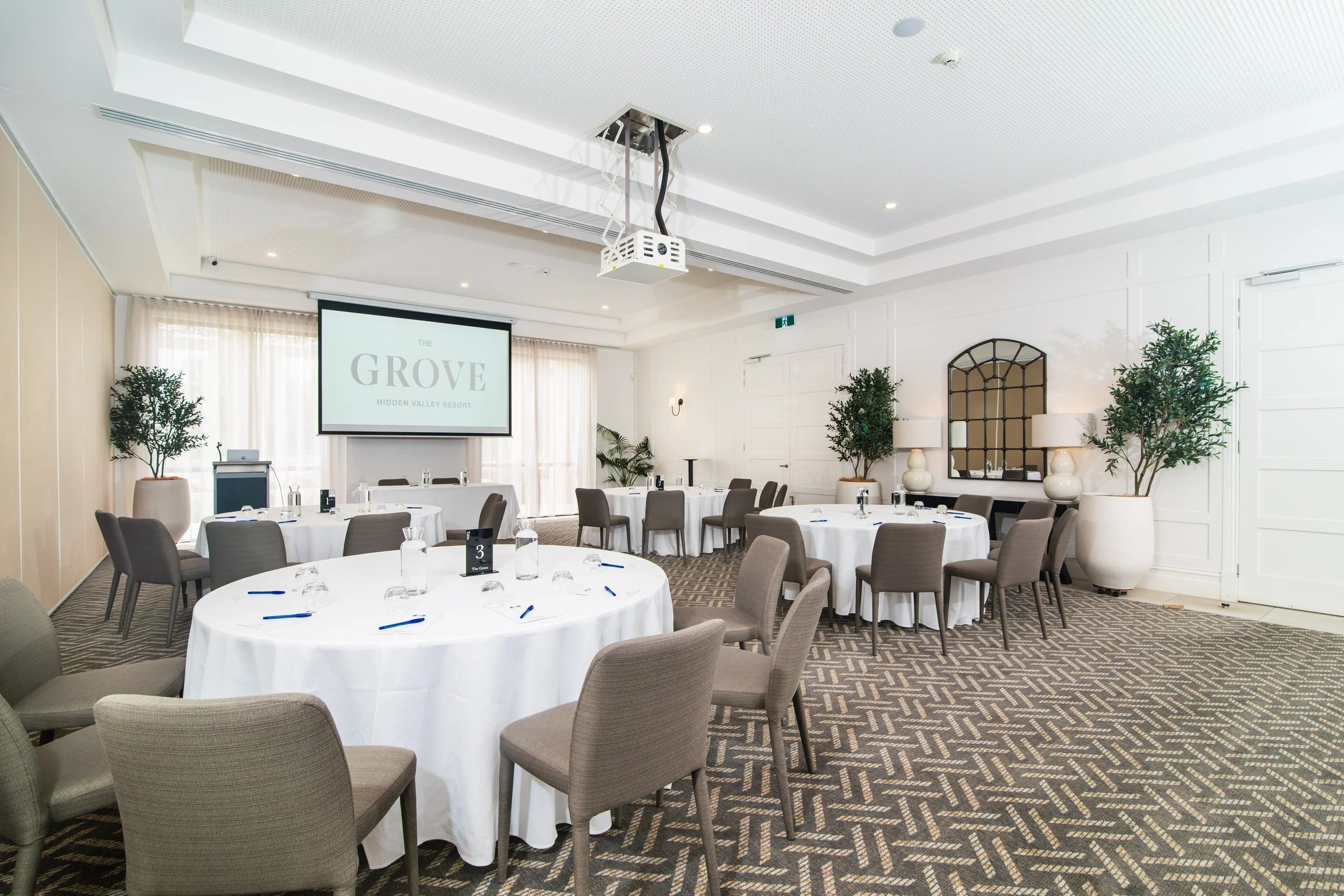 An empty conference room set up with round tables covered in white tablecloths, each with water bottles, glasses, and pens. Large potted plants and a decorative mirror decorate the white walls. A large screen displays 'The Grove Hidden Valley Resort'