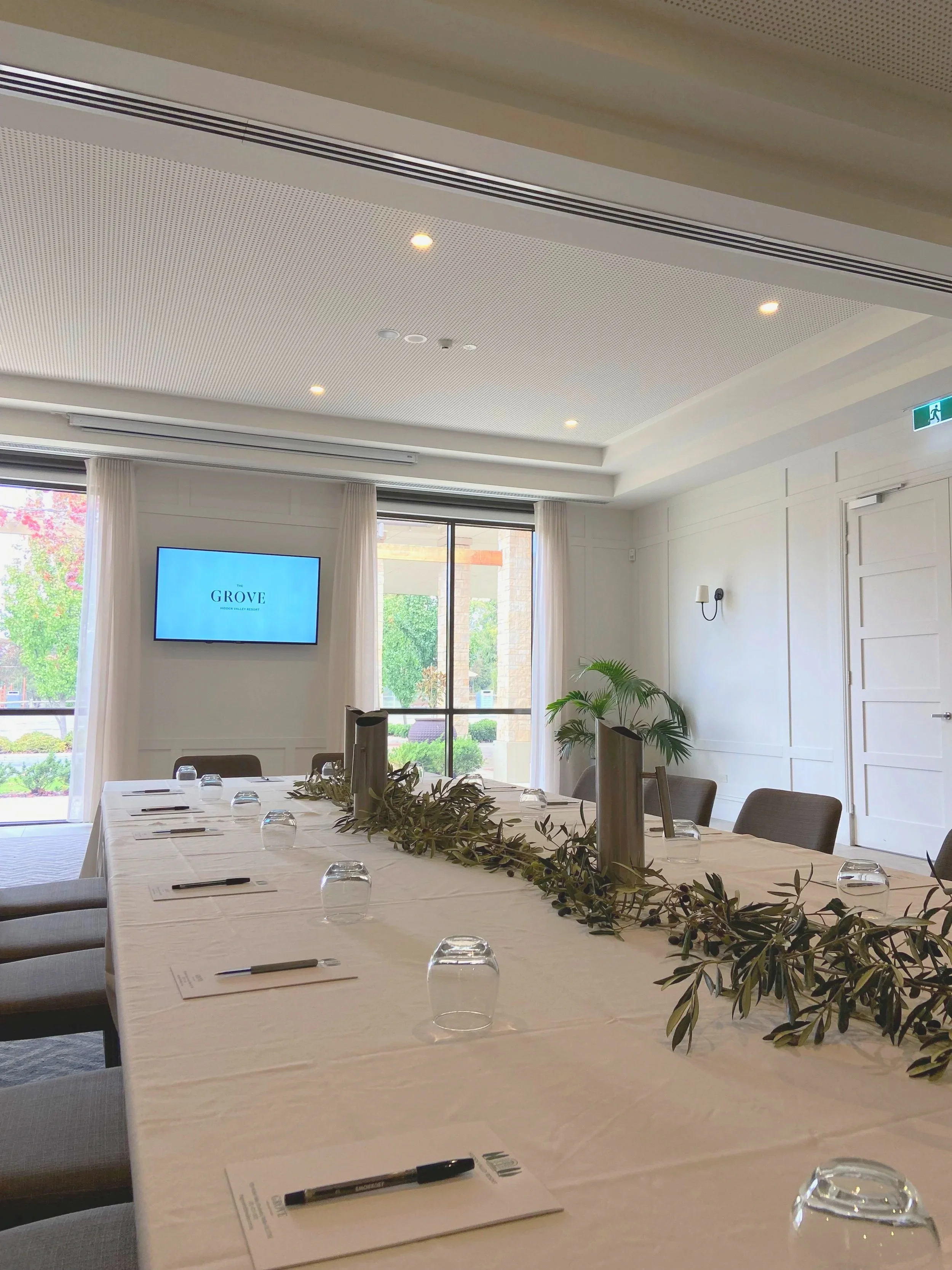 Conference room with a long table set with notepads, pens, upside-down glasses, and a decorative greenery runner, with a large window showing trees outside and a wall-mounted TV displaying 'The Grove' logo.