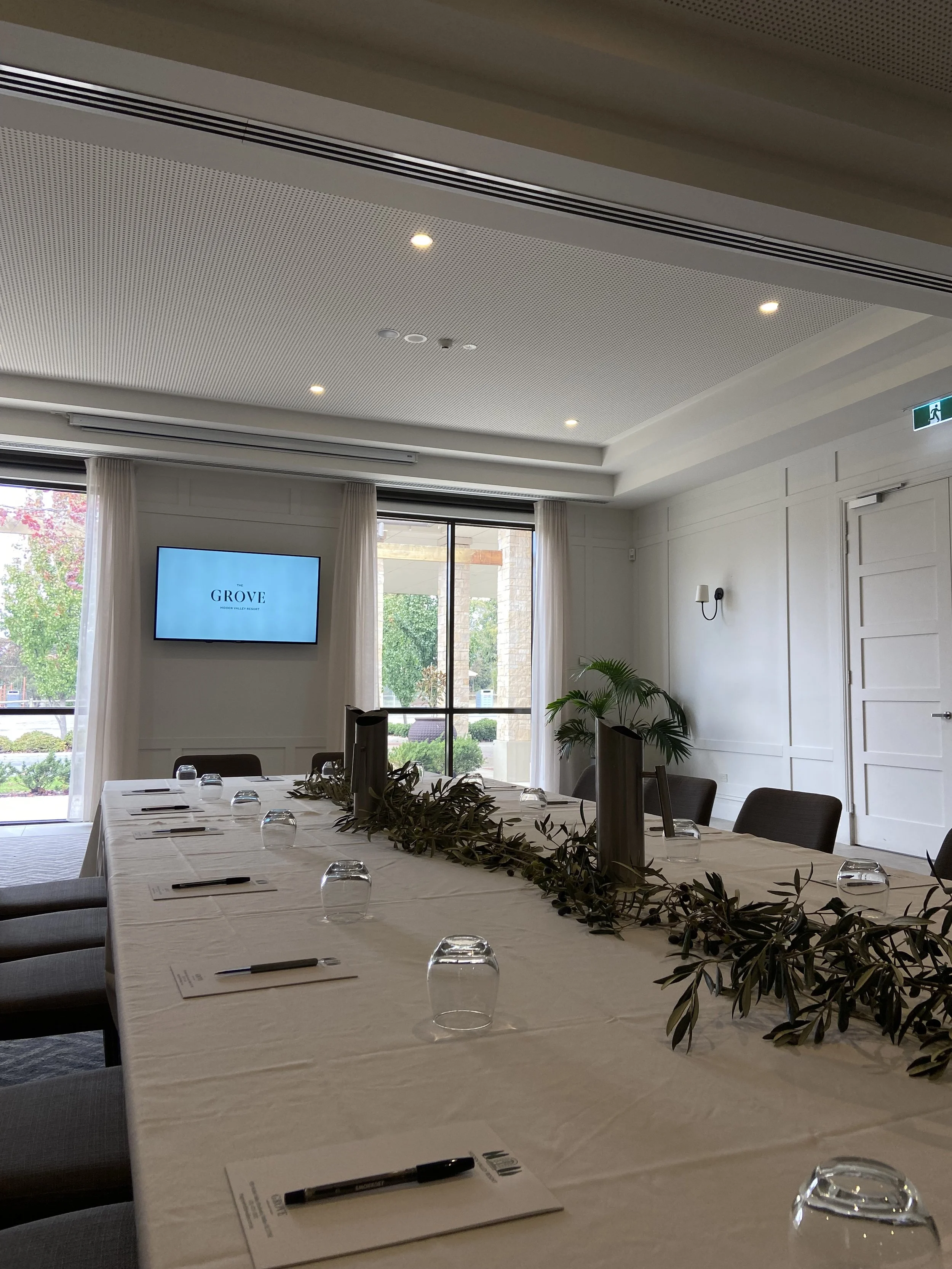 A conference room with a long table set with notepads, pens, upside-down glasses, and a leafy centerpiece. The room features white walls, large windows with white curtains, and a mounted TV displaying the logo 'The Grove'.