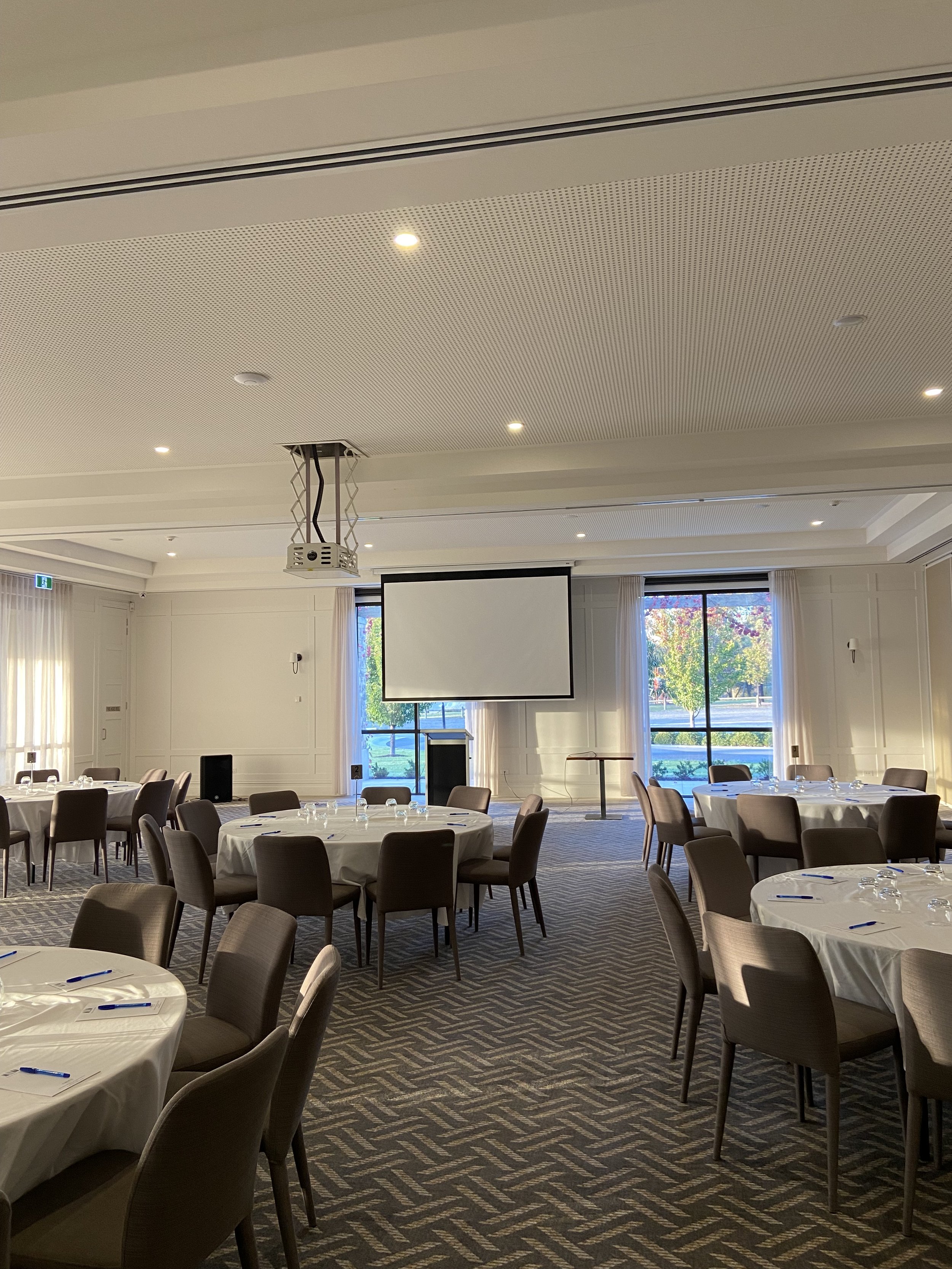 A banquet room with round tables covered in white tablecloths, set with glasses and notepads, arranged for a meeting or event. There is a large projection screen hanging from the ceiling in front of tall windows with white curtains, showing a view of trees outside.
