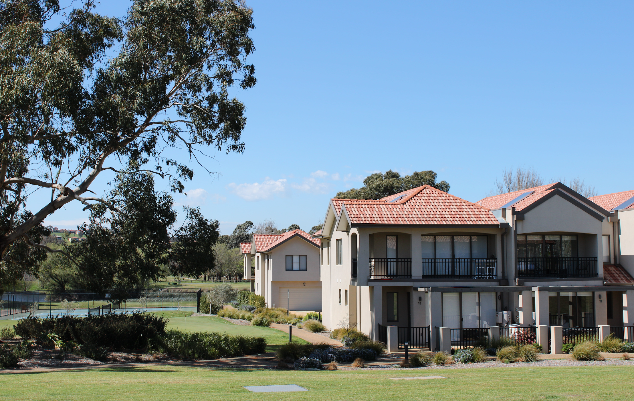 Residential complex with modern white houses, red-tiled roofs, and balconies, set in a landscaped area with grass, pathways, and a tennis court in the background, under a clear blue sky.