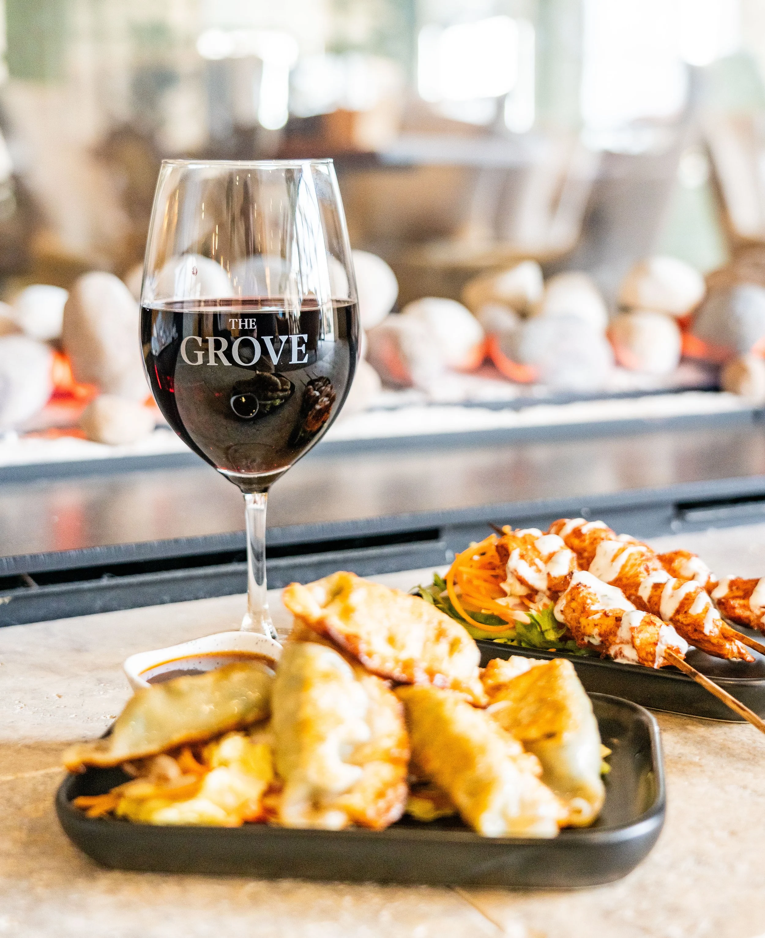 A glass of red wine on a counter next to a black tray with fried appetizers and a plate with skewers of grilled meat and vegetables in a restaurant setting.