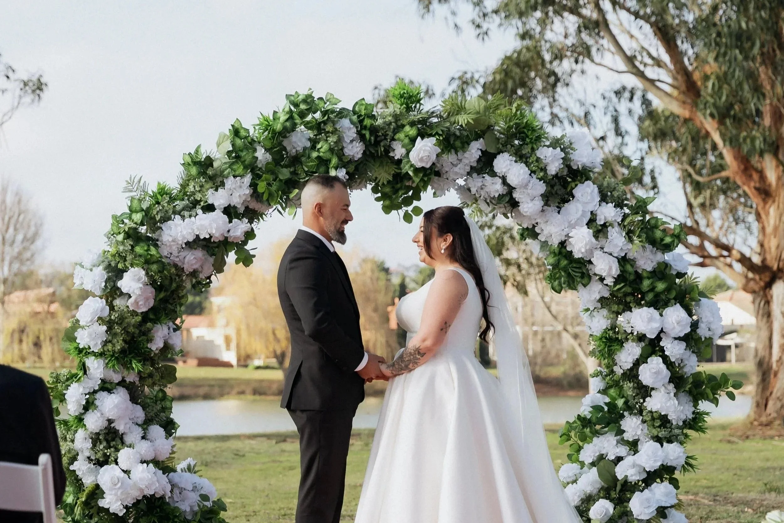 A wedding ceremony with a couple holding hands underneath a floral arch outdoors near a pond, with trees and buildings in the background.