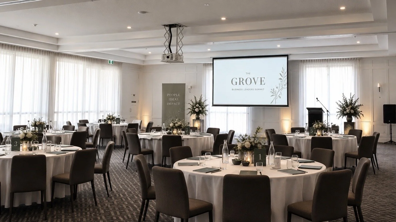 A banquet room with round tables covered in white tablecloths, set with glasses and notepads, arranged for a meeting or event. There is a large projection screen hanging from the ceiling in front of tall windows with white curtains, showing a view of trees outside.