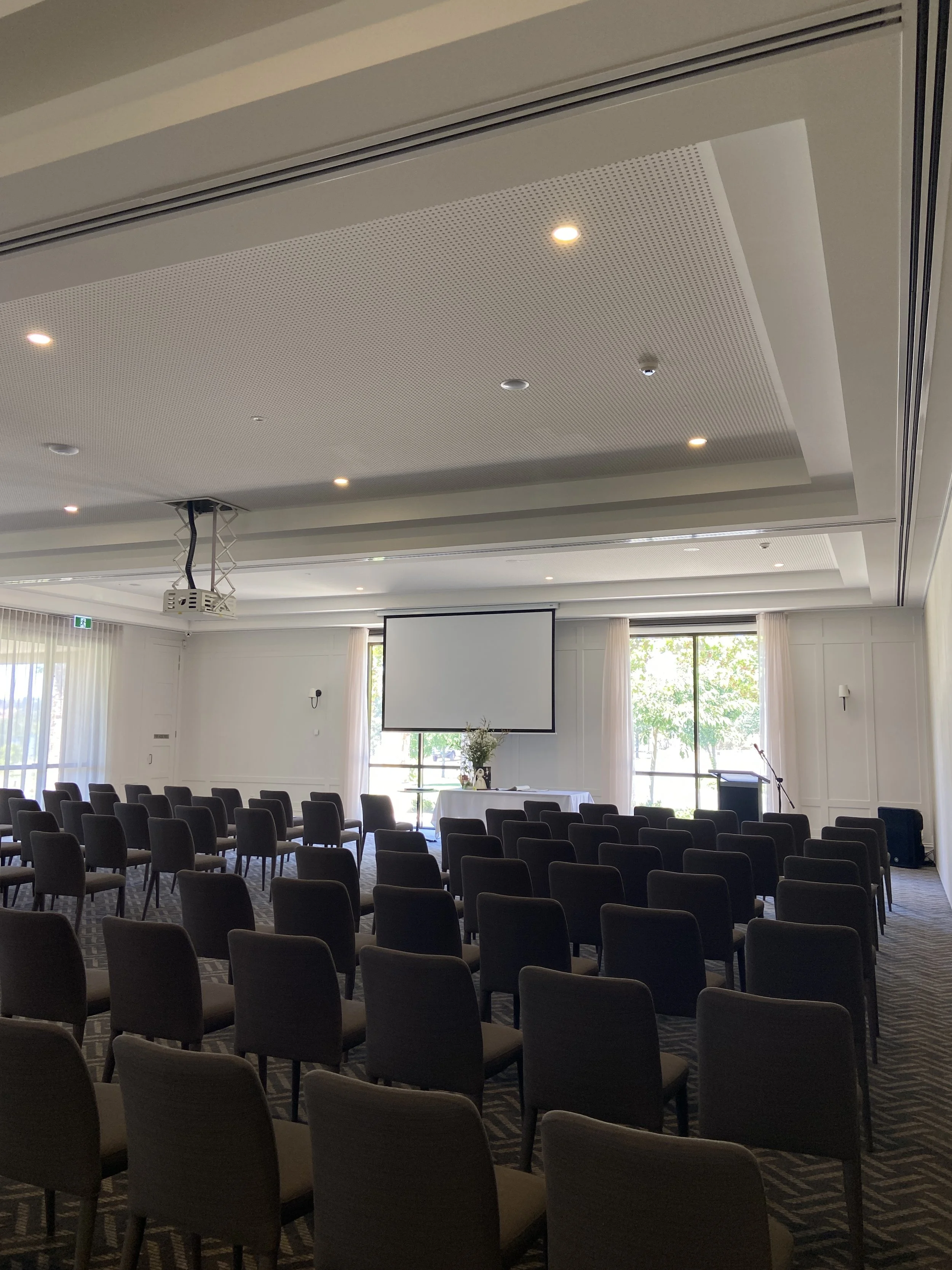 Empty conference room with round tables covered in white tablecloths, chairs, a large projector screen, and large windows with curtains showing trees outside.
