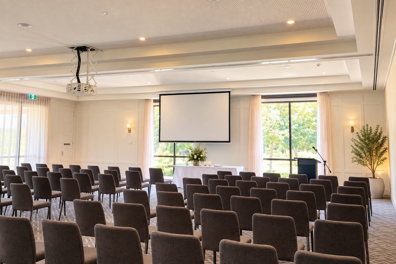Empty conference room with round tables covered in white tablecloths, chairs, a large projector screen, and large windows with curtains showing trees outside.