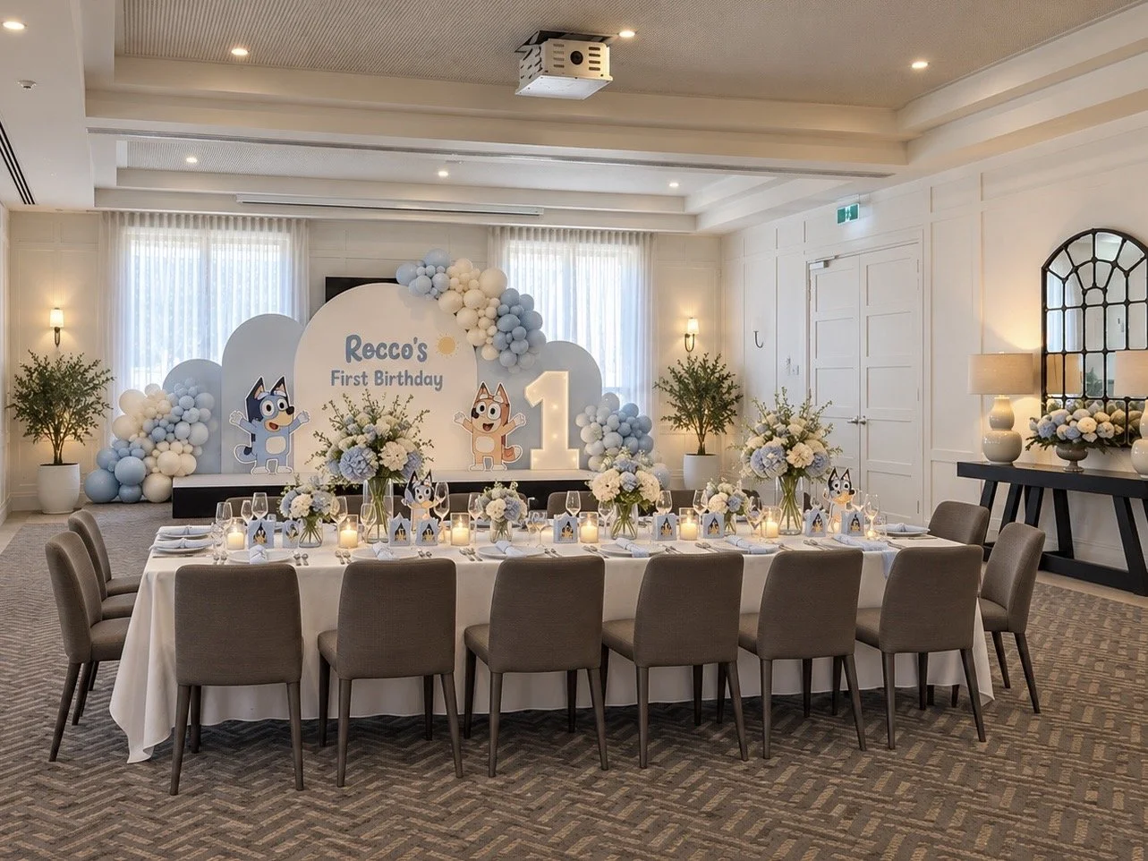 Round banquet table set with wine glasses, water glasses, cutlery, and napkins, in an elegantly decorated room with white paneled walls, lamps, floral arrangements, and a large mirror.