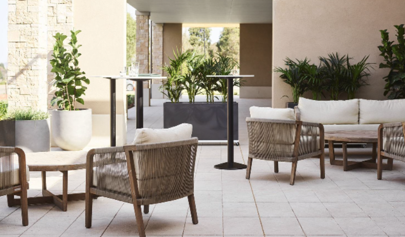 Outdoor patio with wicker chairs, a cushioned sofa, high tables, and potted plants in a modern setting.