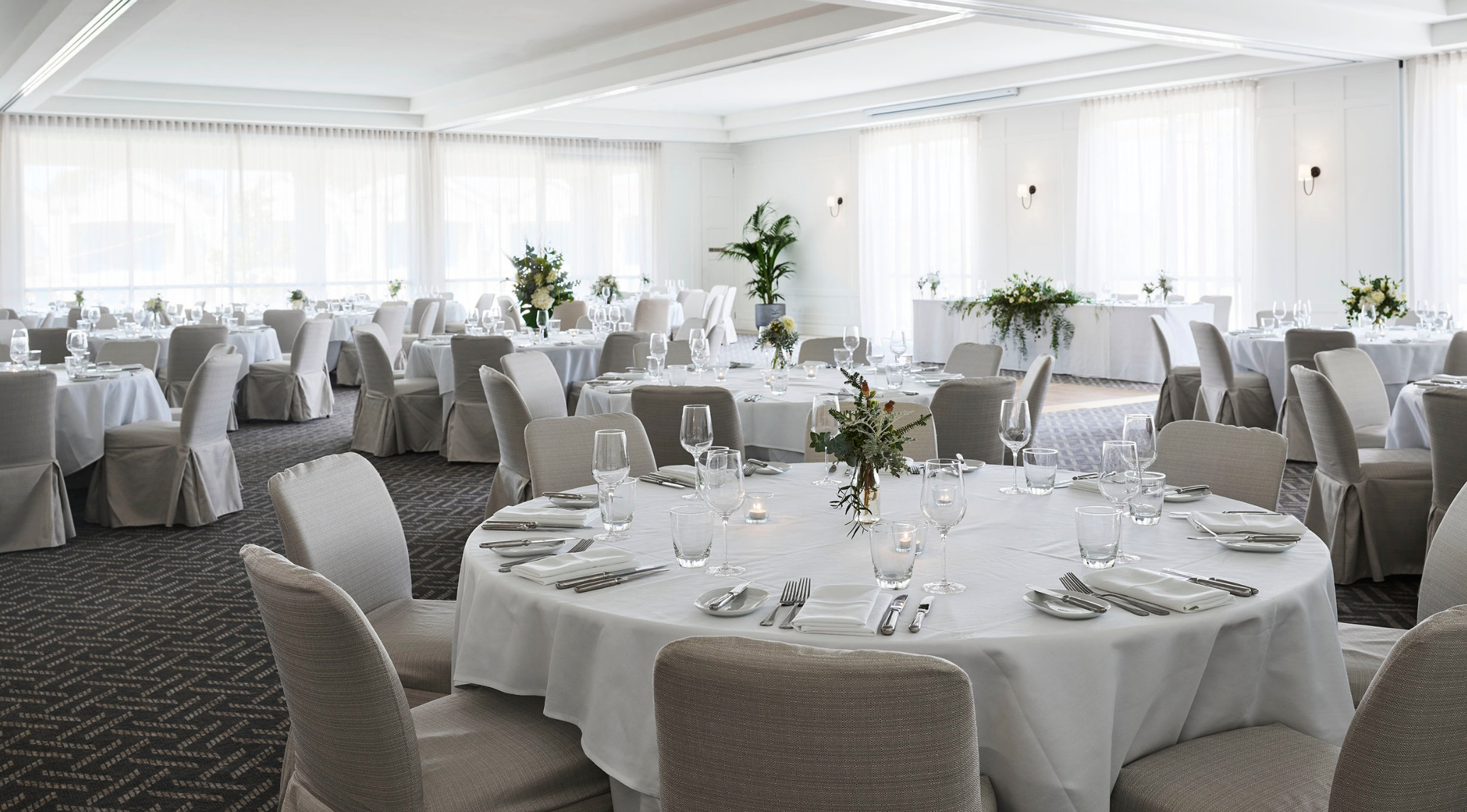 Elegant banquet hall with round tables covered in white tablecloths, set with glassware, silverware, and white napkins, decorated with floral centerpieces, large windows with sheer curtains, and neutral-colored chairs.