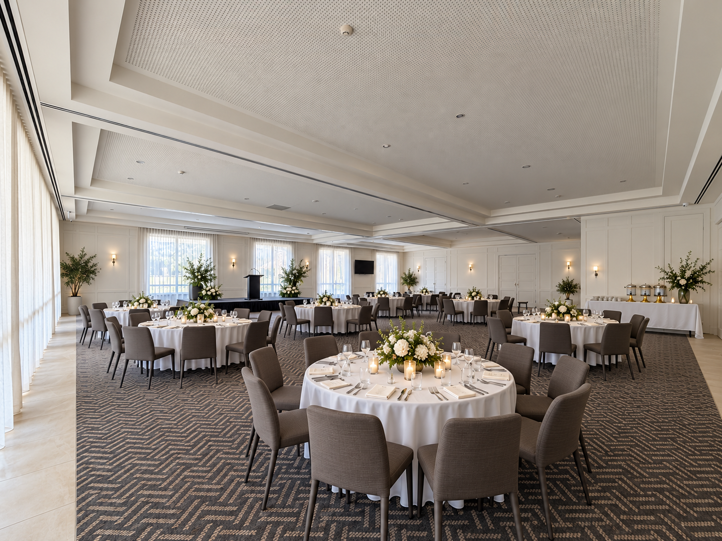 Elegant event space with round tables covered in white cloth, brown chairs, and small green plant centerpieces. Soft natural light filters through sheer curtains, and a flat-screen TV is mounted on the wall. Decor includes vases and lamps, and the ceiling has recessed lighting.