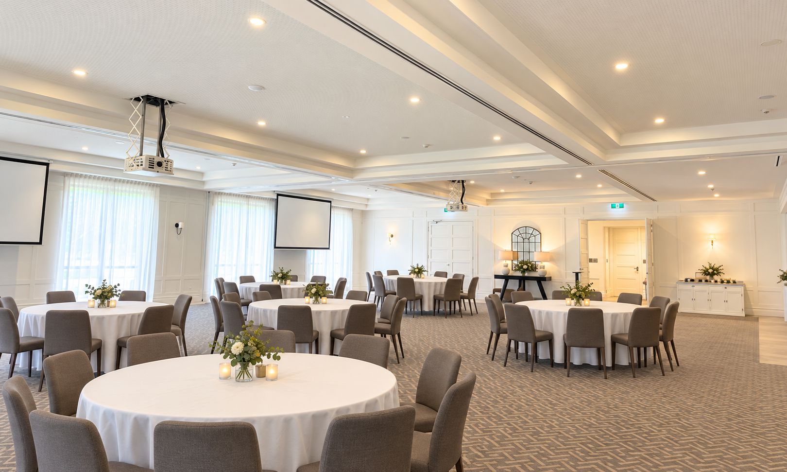 Elegant event space with round tables covered in white cloth, brown chairs, and small green plant centerpieces. Soft natural light filters through sheer curtains, and a flat-screen TV is mounted on the wall. Decor includes vases and lamps, and the ceiling has recessed lighting.