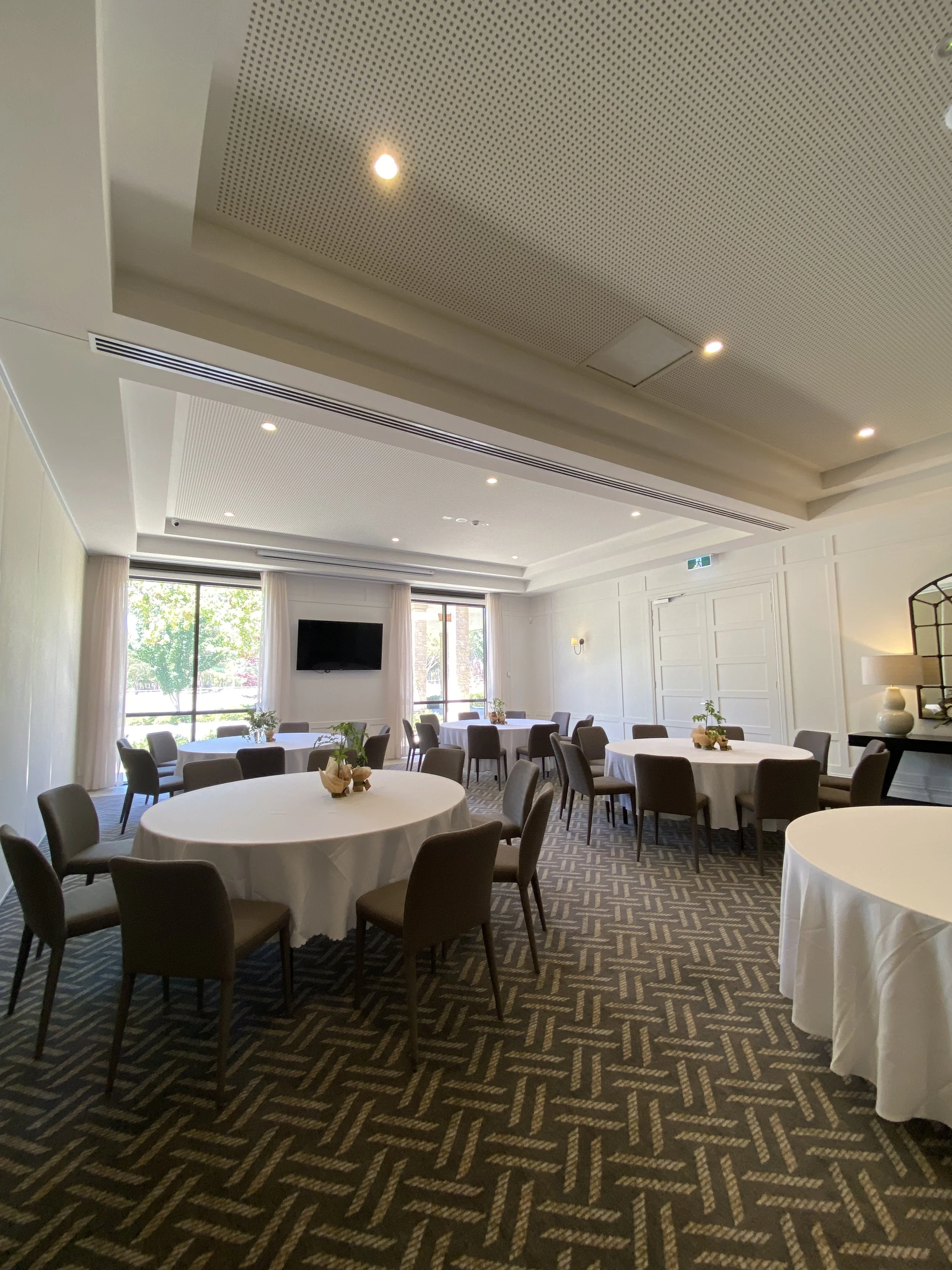 Round banquet table set with wine glasses, water glasses, cutlery, and napkins, in an elegantly decorated room with white paneled walls, lamps, floral arrangements, and a large mirror.