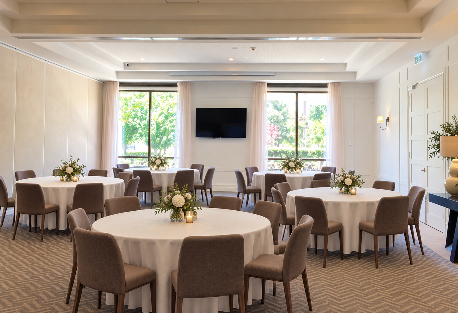 Round banquet table set with wine glasses, water glasses, cutlery, and napkins, in an elegantly decorated room with white paneled walls, lamps, floral arrangements, and a large mirror.