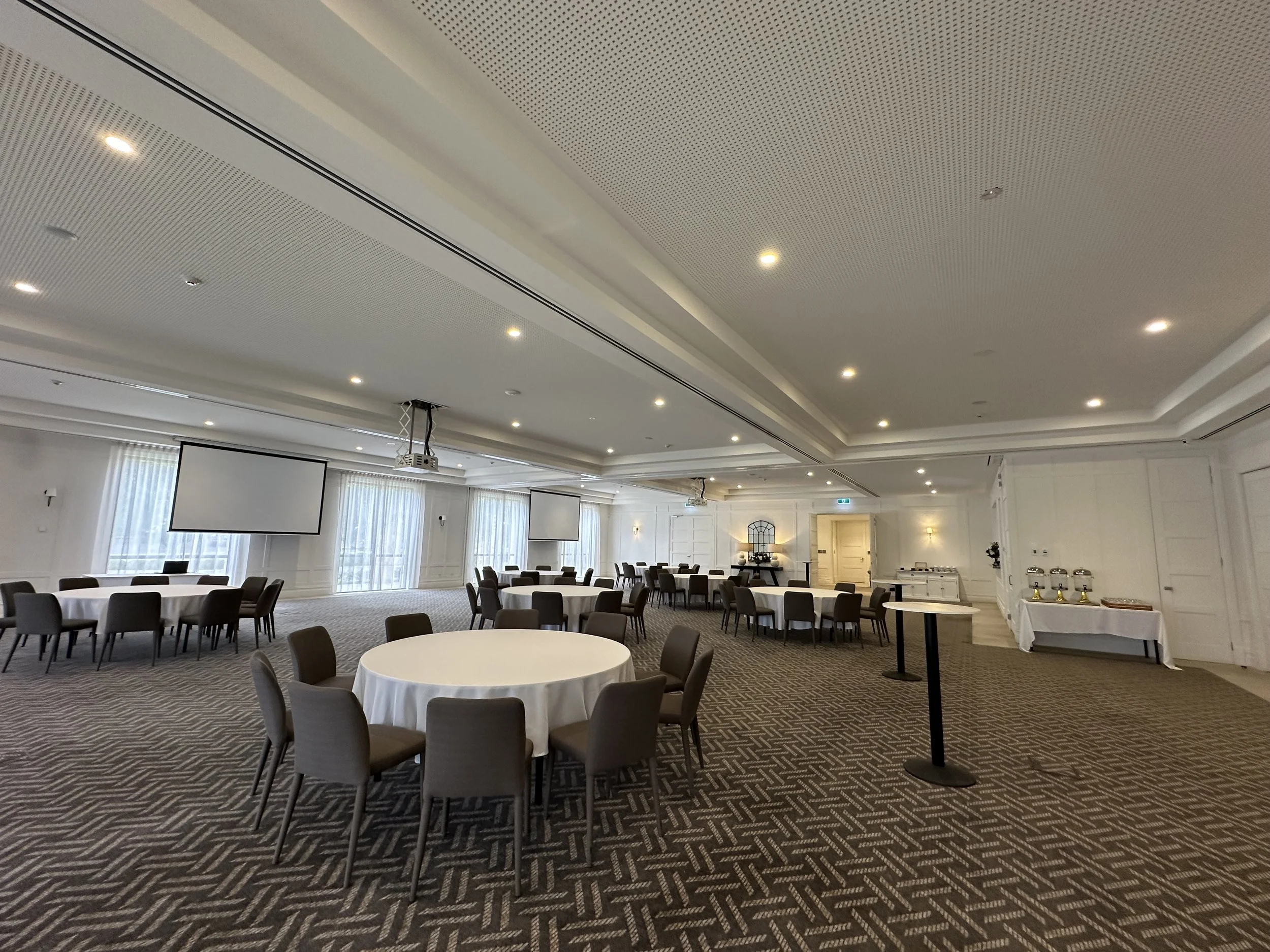 Elegant event space with round tables covered in white cloth, brown chairs, and small green plant centerpieces. Soft natural light filters through sheer curtains, and a flat-screen TV is mounted on the wall. Decor includes vases and lamps, and the ceiling has recessed lighting.