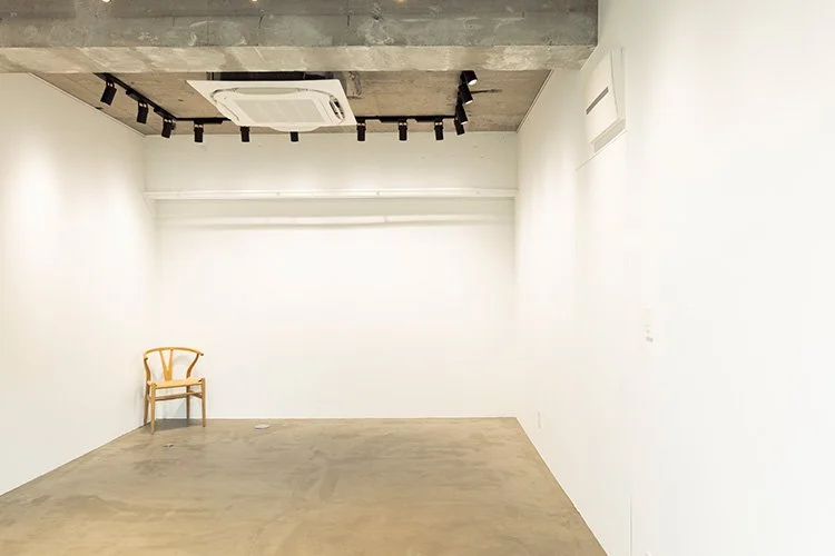 白い壁の部屋の左側に木製の椅子が置かれている。天井には空調とスポットライトが設置されている。