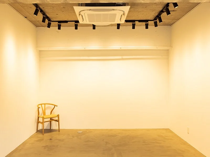 白い壁のシンプルな部屋に一脚の木製椅子が左側に置かれている。天井には照明とエアコンダクトが見える。