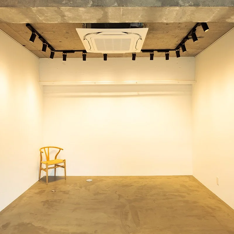 白い壁のシンプルな部屋に一脚の木製椅子が左側に置かれている。天井には照明とエアコンダクトが見える。