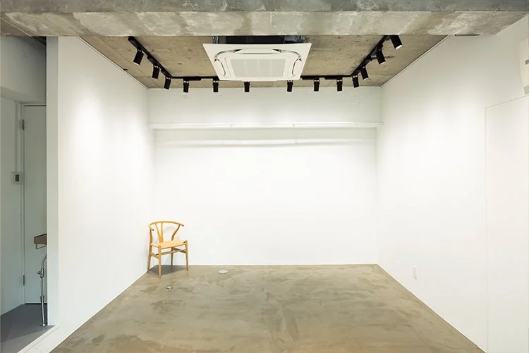 白い壁の部屋の左側に木製の椅子が置かれている。天井には空調とスポットライトが設置されている。