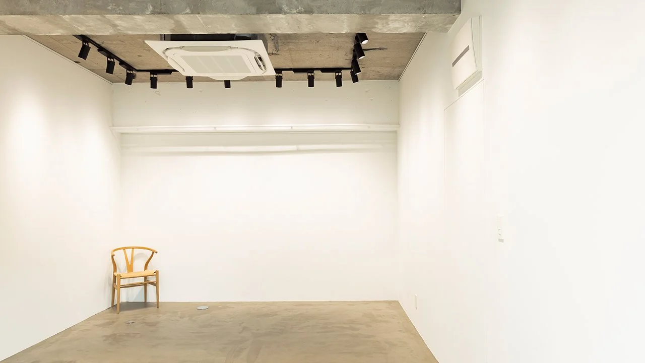 白い壁の部屋に木製の椅子とエアコンと照明があり、シンプルな内装の空間