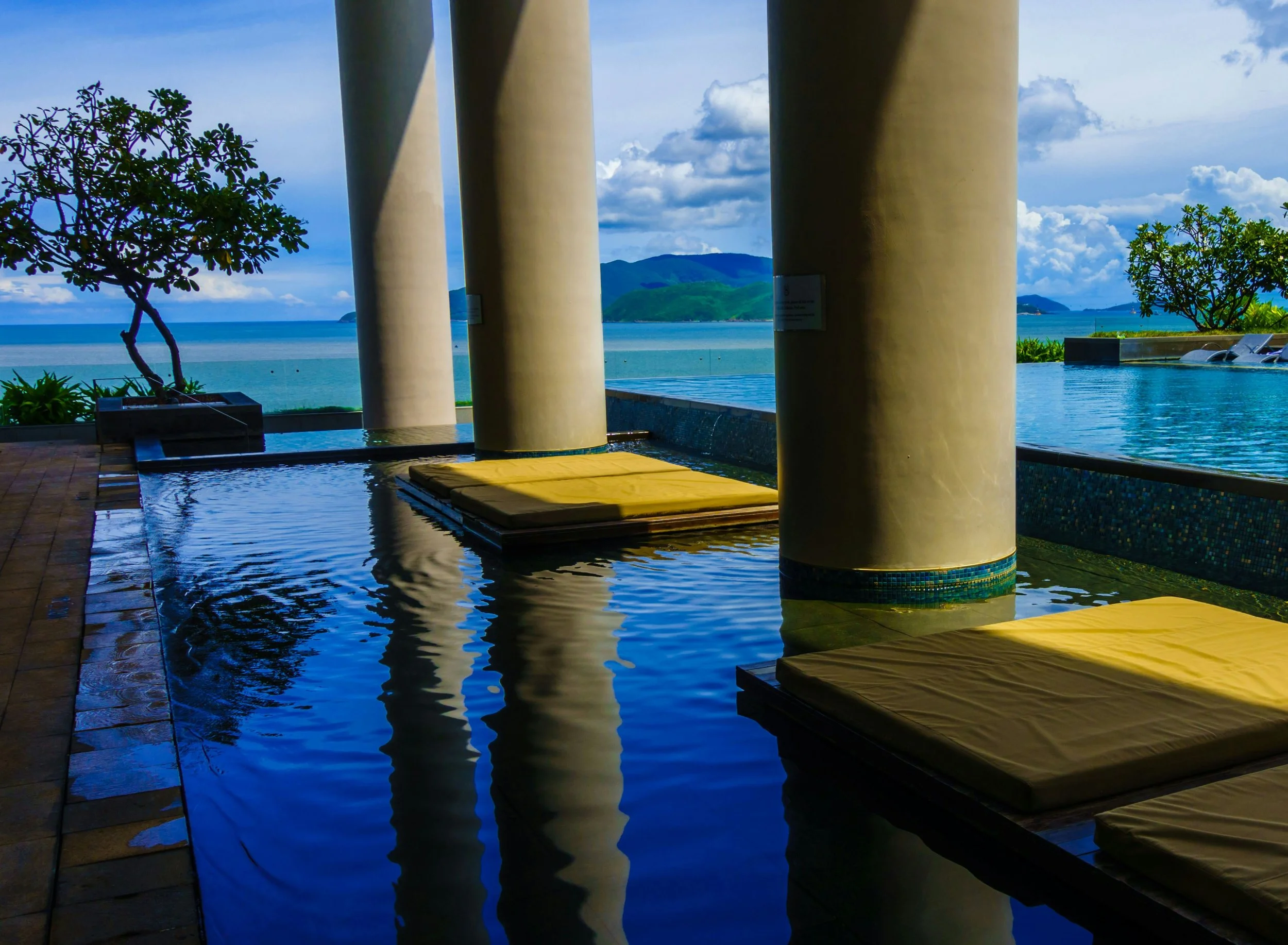 Luxury poolside area with lounge chairs, large beige pillars, and a scenic ocean view with islands and a partly cloudy sky in the background.