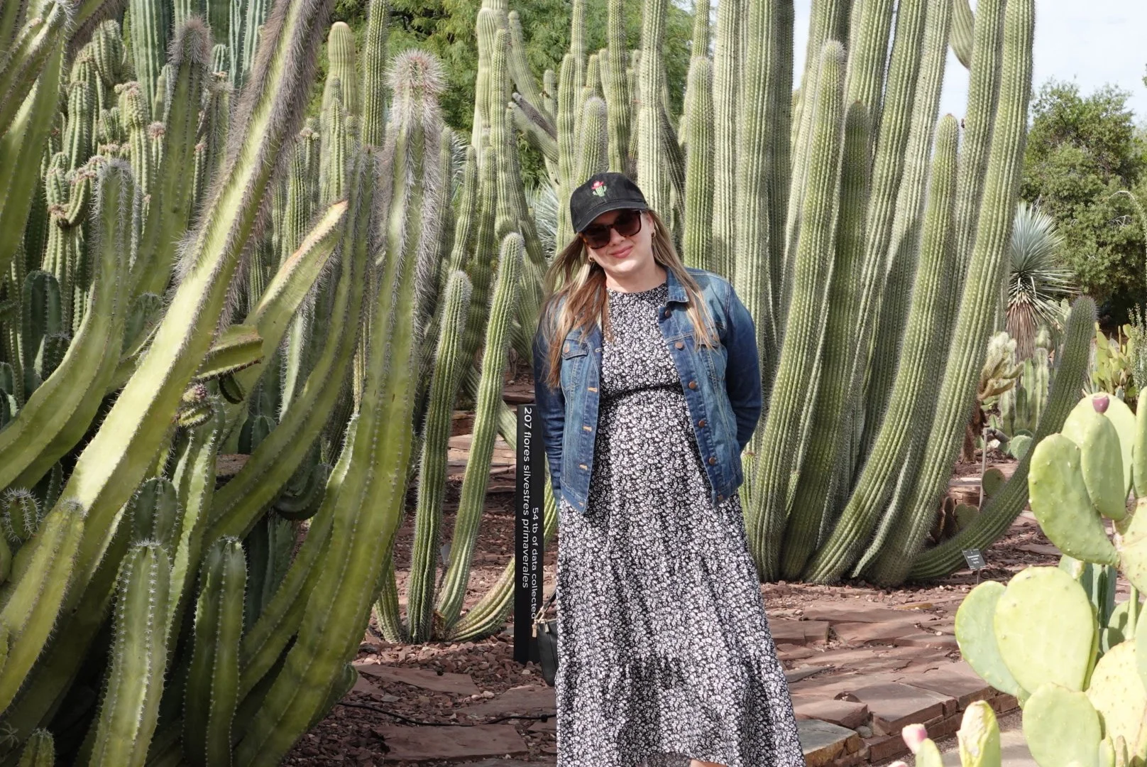 A woman standing amid tall cacti, wearing a black cap, sunglasses, a denim jacket, and a long floral dress.
