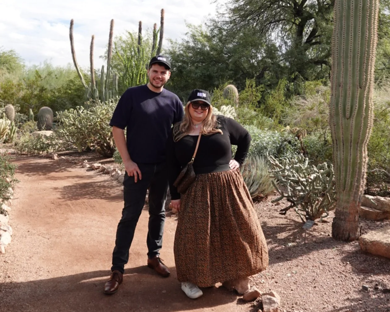 Two people standing on a dirt path in a desert botanical garden, surrounded by cacti and desert plants, with a partly cloudy sky above.
