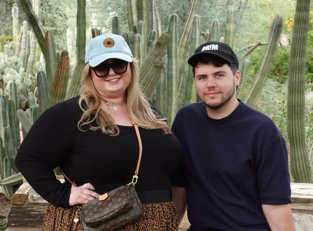 A woman and a man are standing outdoors in front of tall cacti. The woman is wearing a light blue cap, sunglasses, a black long-sleeve top, and leopard print pants. The man is wearing a black cap and a navy blue t-shirt. They are smiling and posing for the photo.