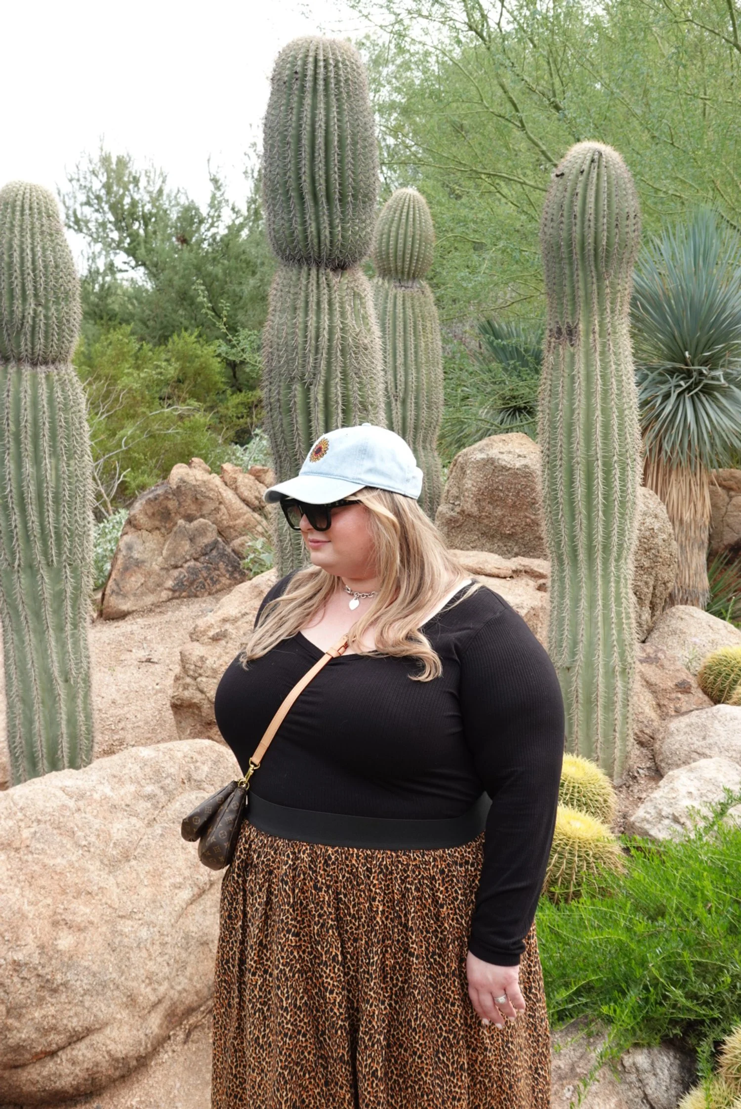 A woman standing in front of tall cacti and desert plants, wearing a black top, leopard-print skirt, baseball cap, sunglasses, and a small crossbody purse.