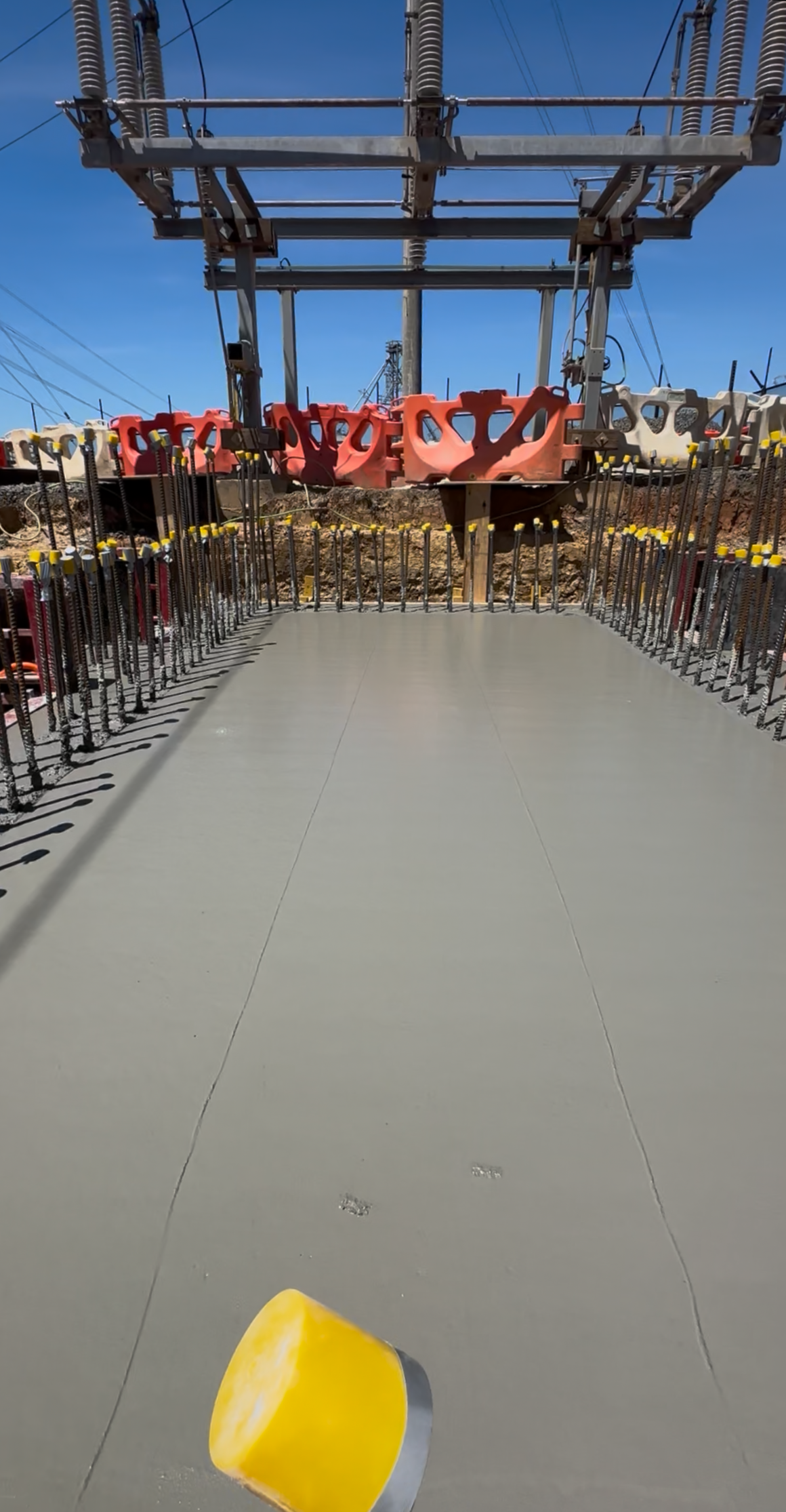 View from a construction worker's perspective on a road with a yellow bucket in the foreground, metal rebar on either side, and electrical infrastructure overhead against a clear blue sky.