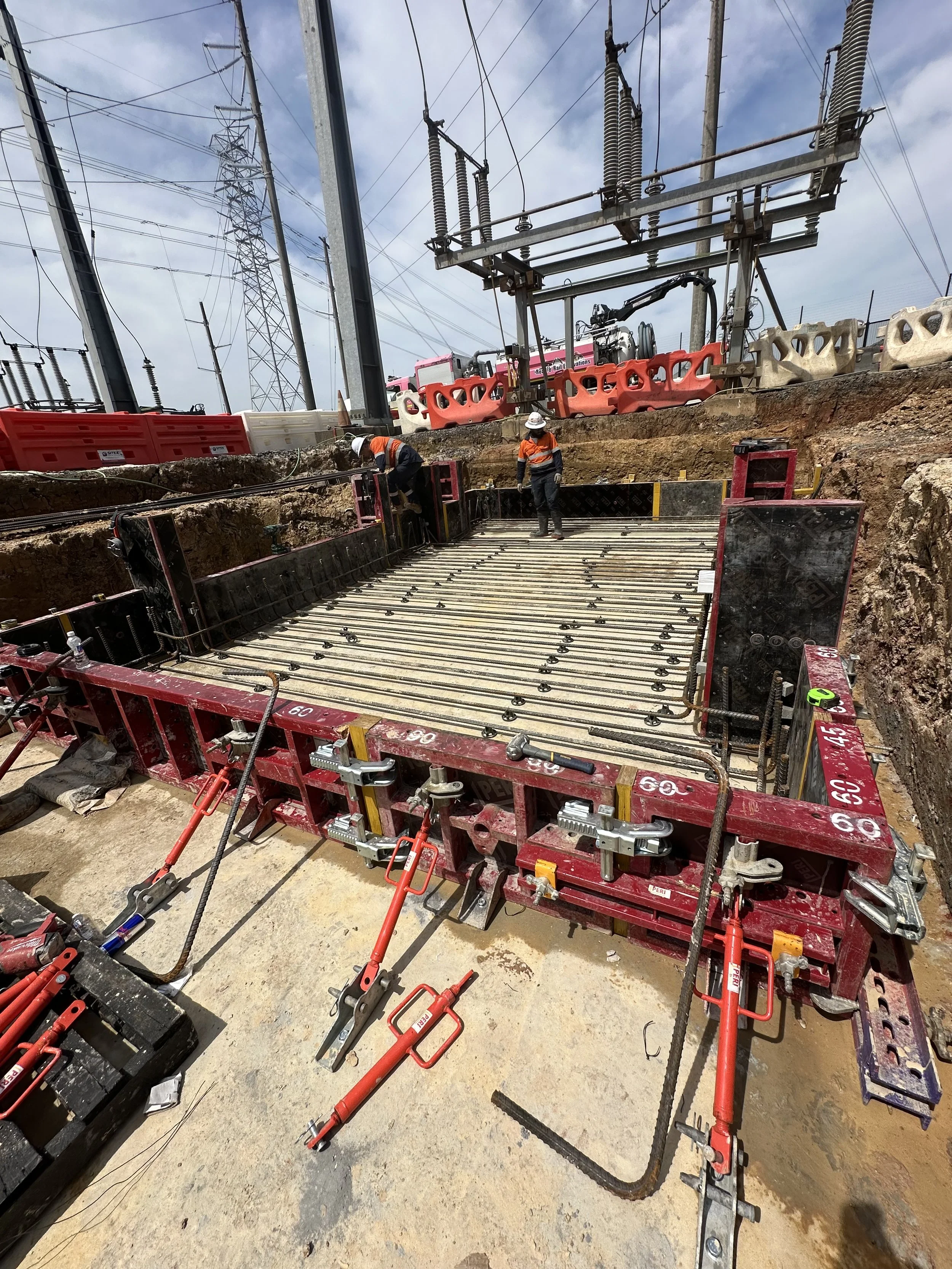 Construction site with workers installing a large concrete form, surrounded by construction tools and equipment, with power lines and transformers in the background.
