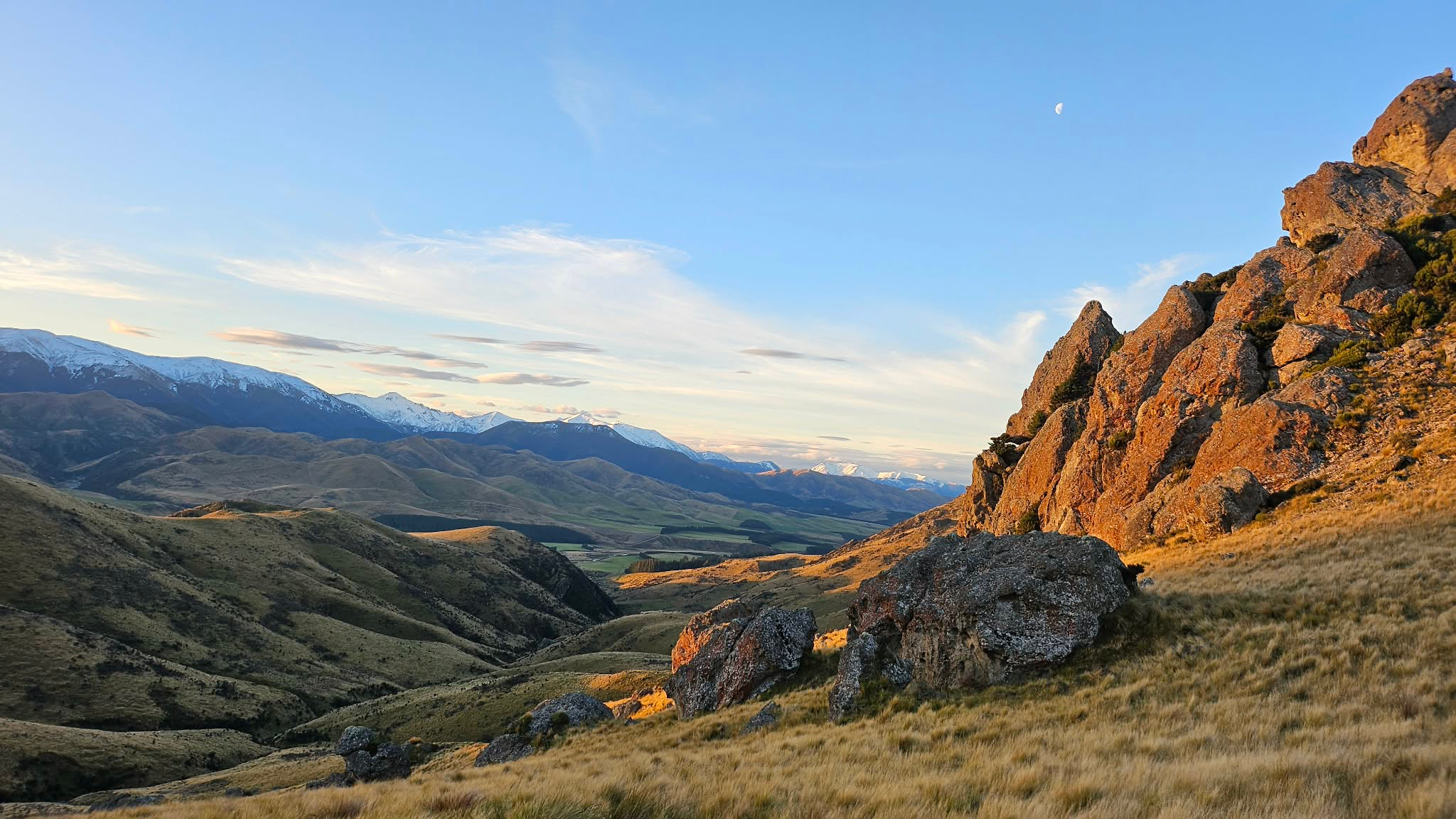 A scenic landscape of rolling hills and mountains with a clear blue sky, some snow-capped peaks, and a visible moon.
