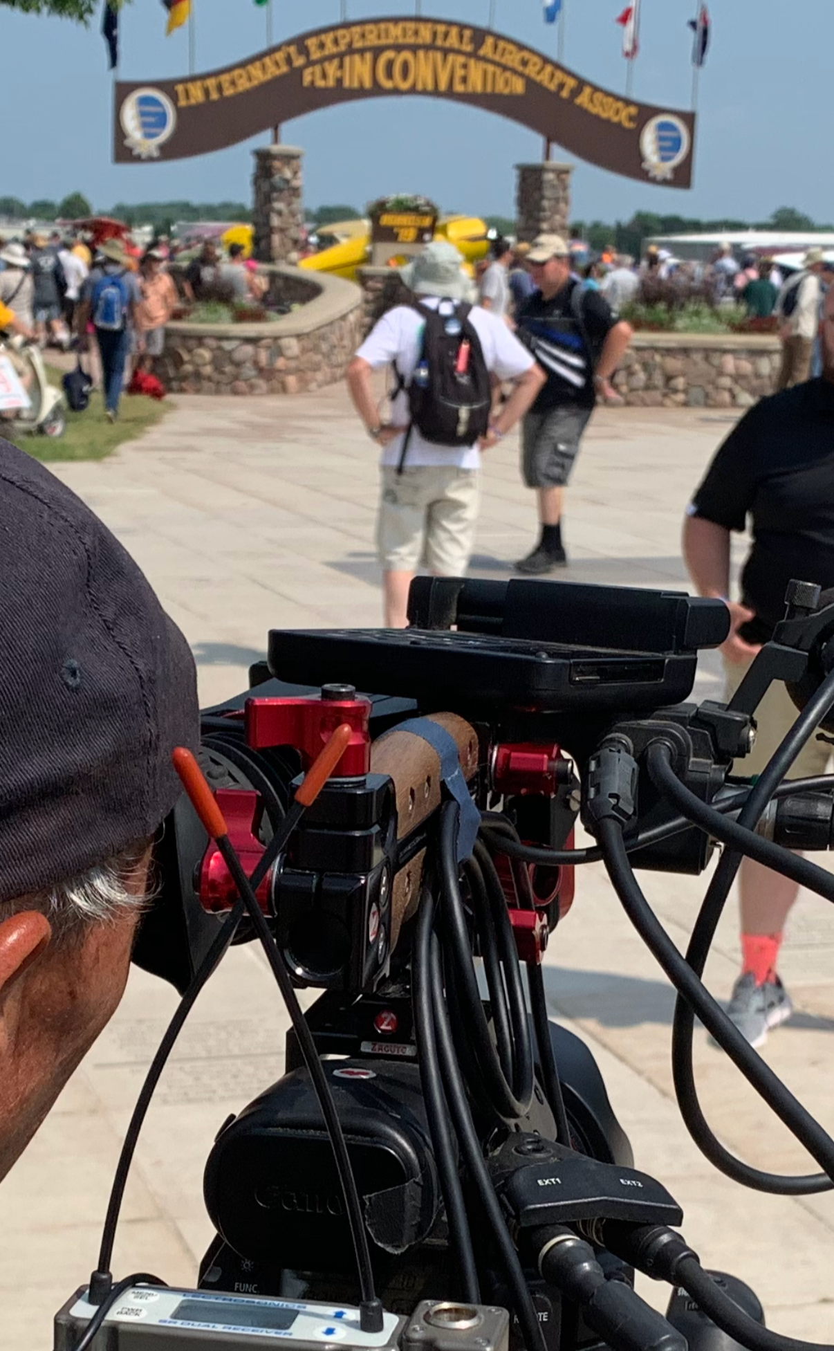 Camera shot focusing on a film or video camera set up at an outdoor event, with the background showing the entrance to a convention with a sign reading 'International Experimental Aircraft Association Fly-In Convention' and numerous attendees walking around.