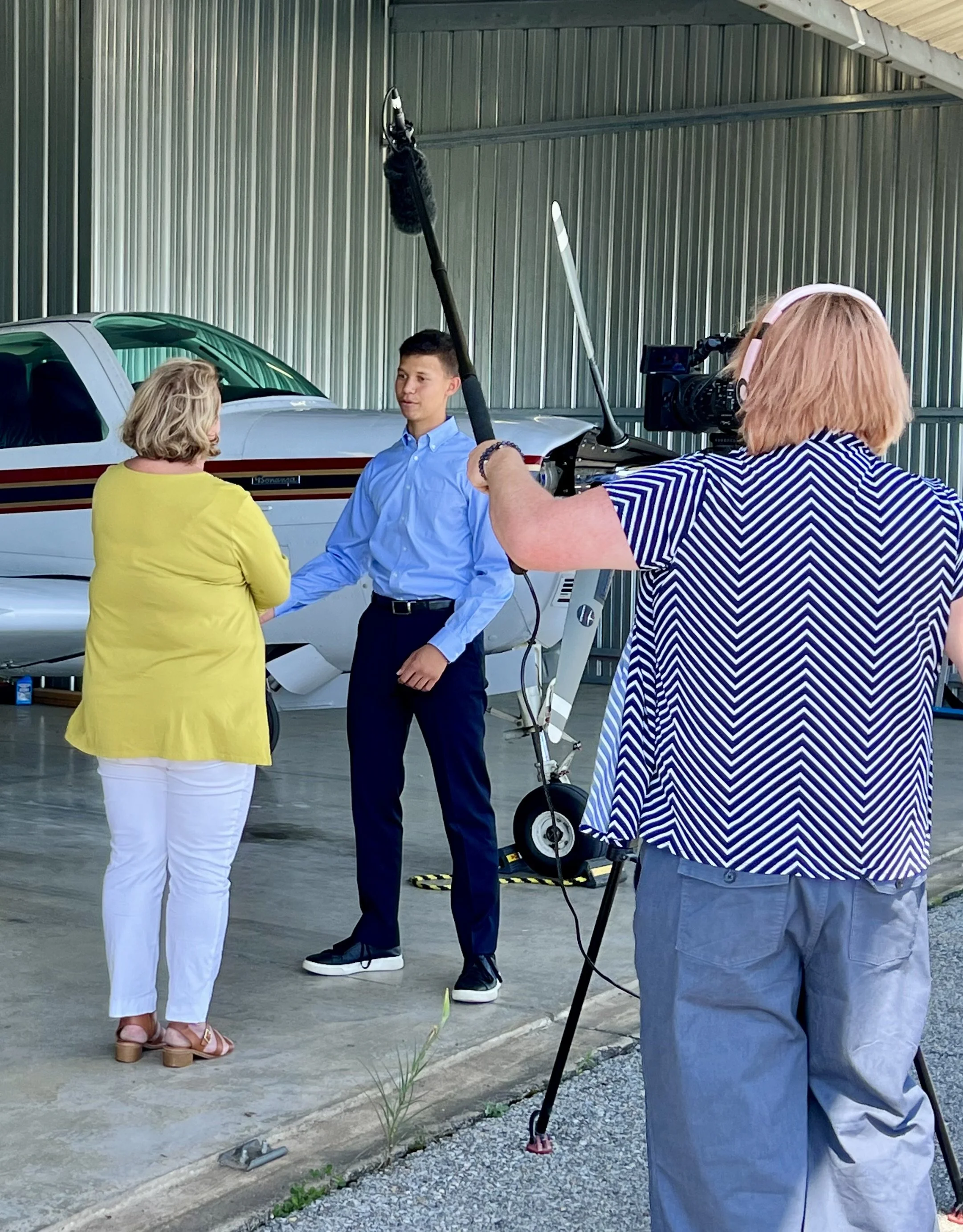 A woman operates a TV camera and a boom microphone while another woman interviews a young man. They are in an open aircraft hangar and an airplane is seen behind the teenager.