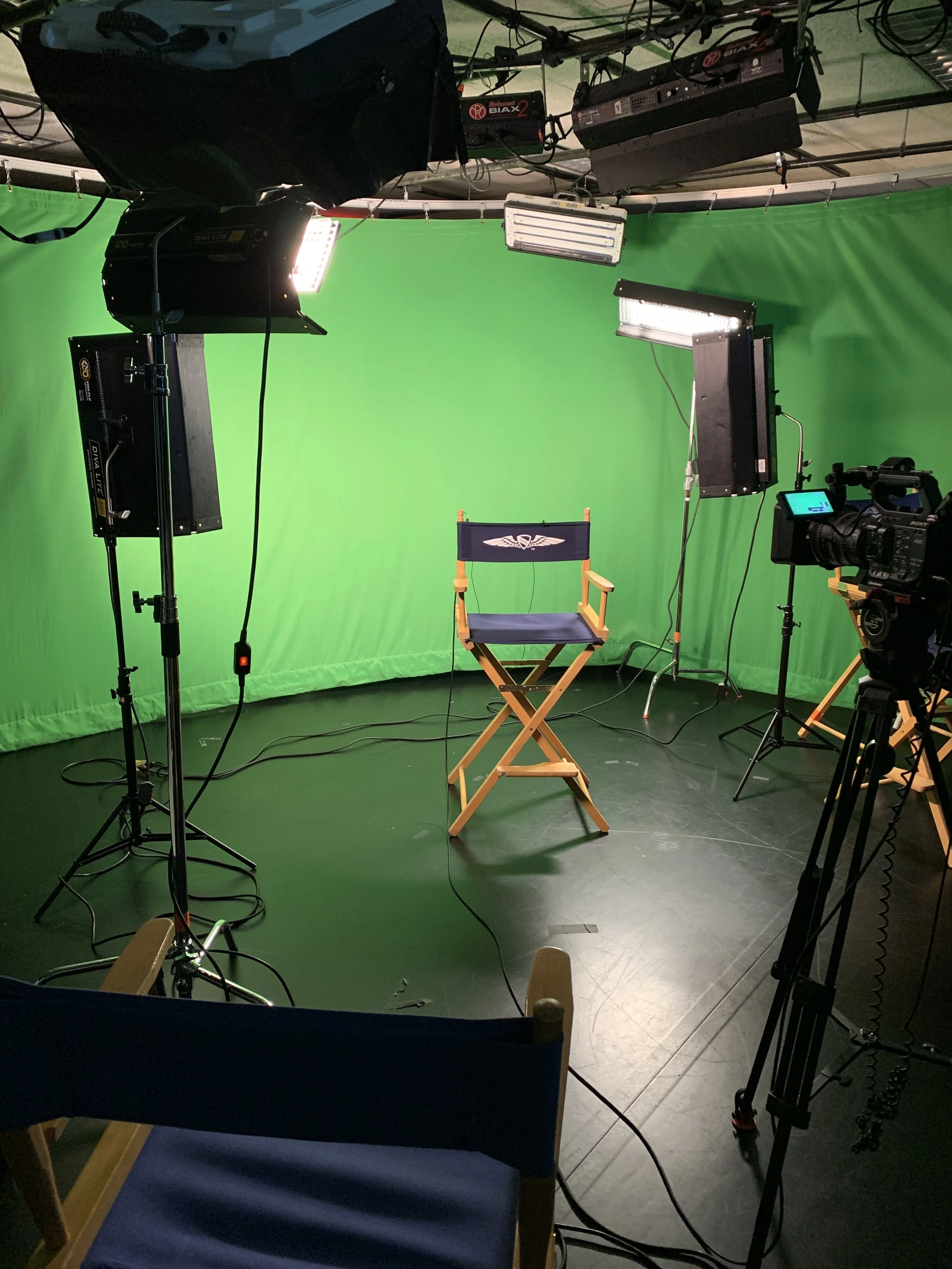 A television studio with lights, cameras, and a green screen background. A directors chair is visible in the shot.